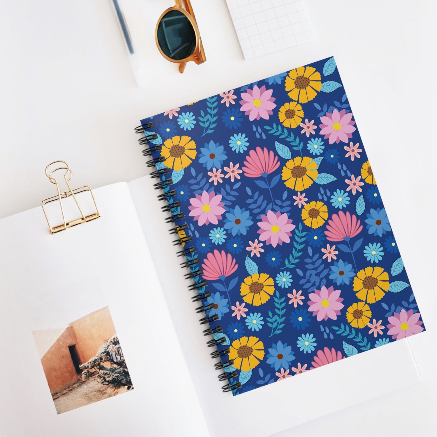 Floral Spiral Notebook with vibrant pink, yellow, and light-blue floral patterns, set within a stylish workspace featuring sunglasses and a grid notepad, embodying FireBrook Designs' joyful and uplifting essence.