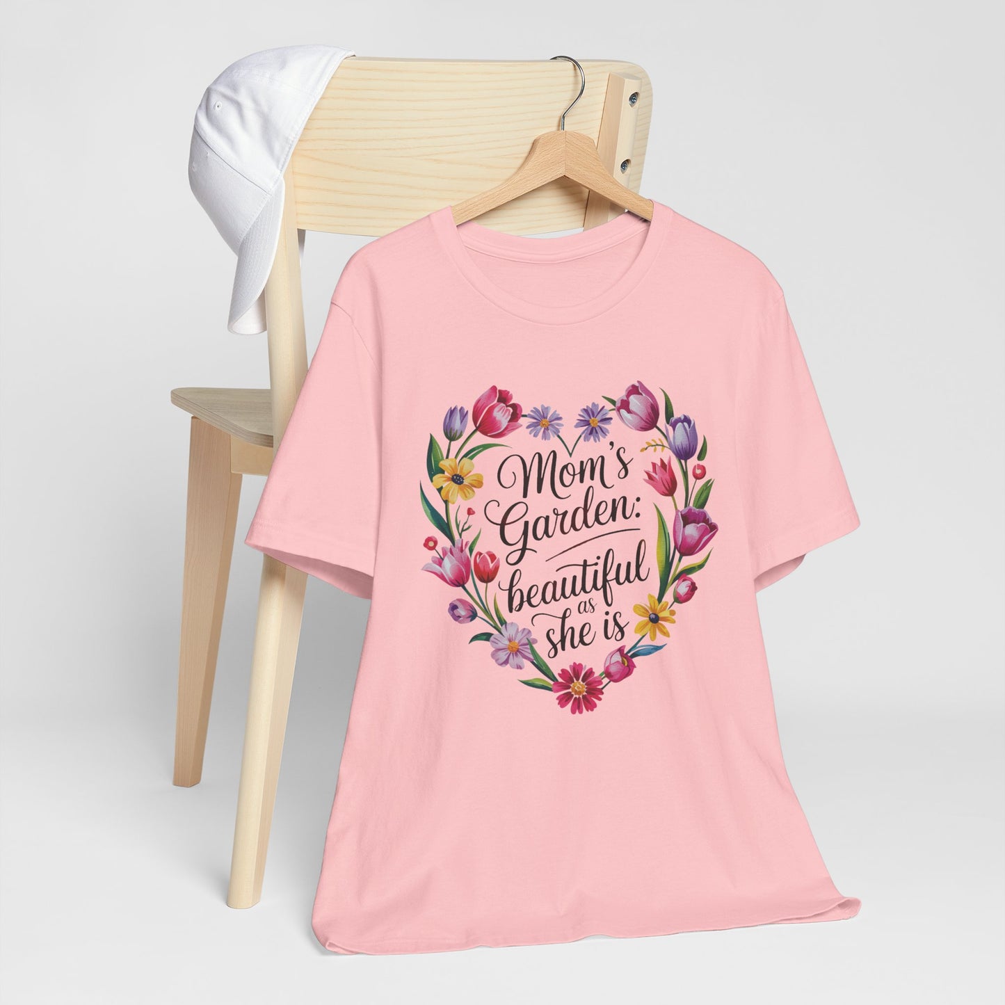 Mom's Garden is as Beautiful as She Is Mother's Day Comfort Colors Unisex T-Shirt, featuring a heart-shaped floral design, draped over a chair with a white hat.
