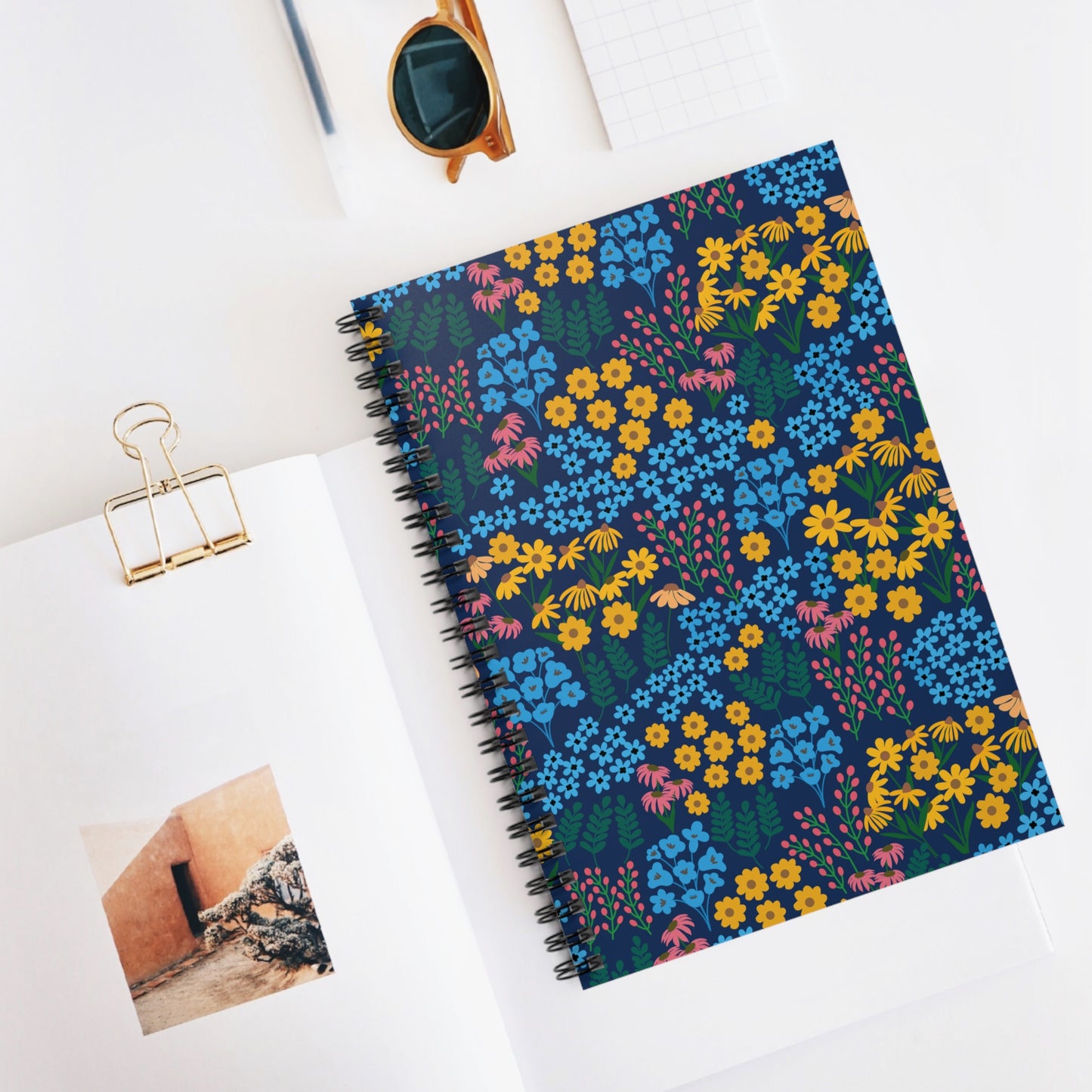 Spiral Notebook, Durable Cover, Rules, 6x8 - Wildflower Meadow Mix, Blue Background, adorned with vibrant floral patterns, rests atop an open book. Nearby are sunglasses, a grid notepad, and a gold binder clip.