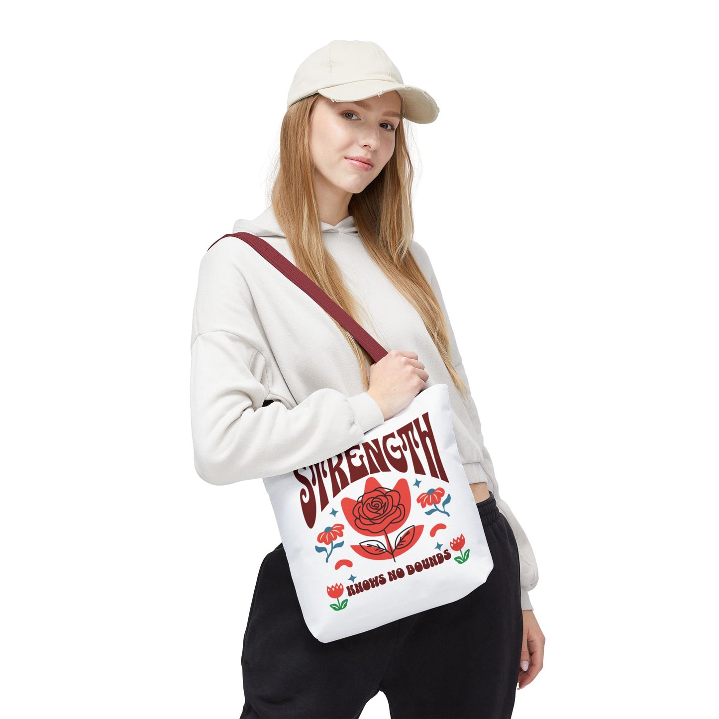 Woman in white sweatshirt and cap holding a Strength Knows No Bounds tote bag, featuring a flower design, showcasing durability and style from FireBrook Essentials.
