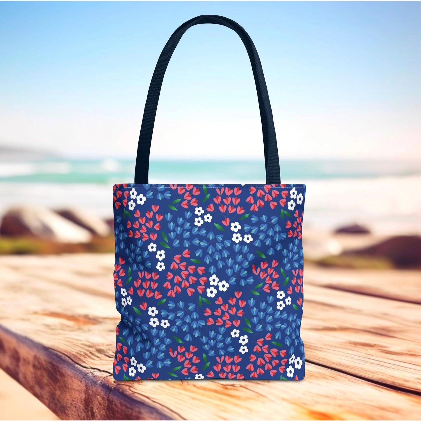 Floral Tote Bag with black handles, featuring vibrant red and blue leafy clusters and tiny white flowers on a dark blue background, standing upright on a wooden surface, ideal for joyful summer adventures.