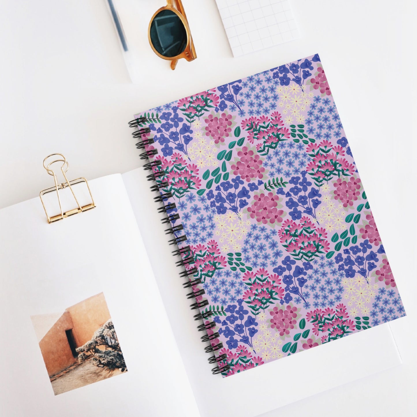 Floral Spiral Notebook with pastel purple-lavender cover featuring vibrant floral clusters, positioned on an open book with a gold binder clip, round sunglasses, and graph paper, embodies FireBrook Designs' joyful, creative essence.