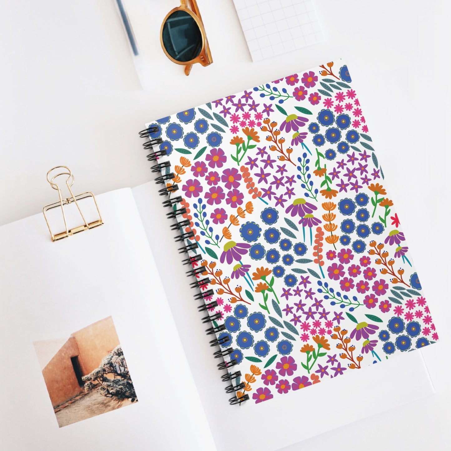 Floral Spiral Notebook with a vibrant cover, surrounded by tortoiseshell sunglasses, graph paper, and a book featuring adobe architecture, embodies FireBrook Essentials' blend of creativity and joy in everyday items.