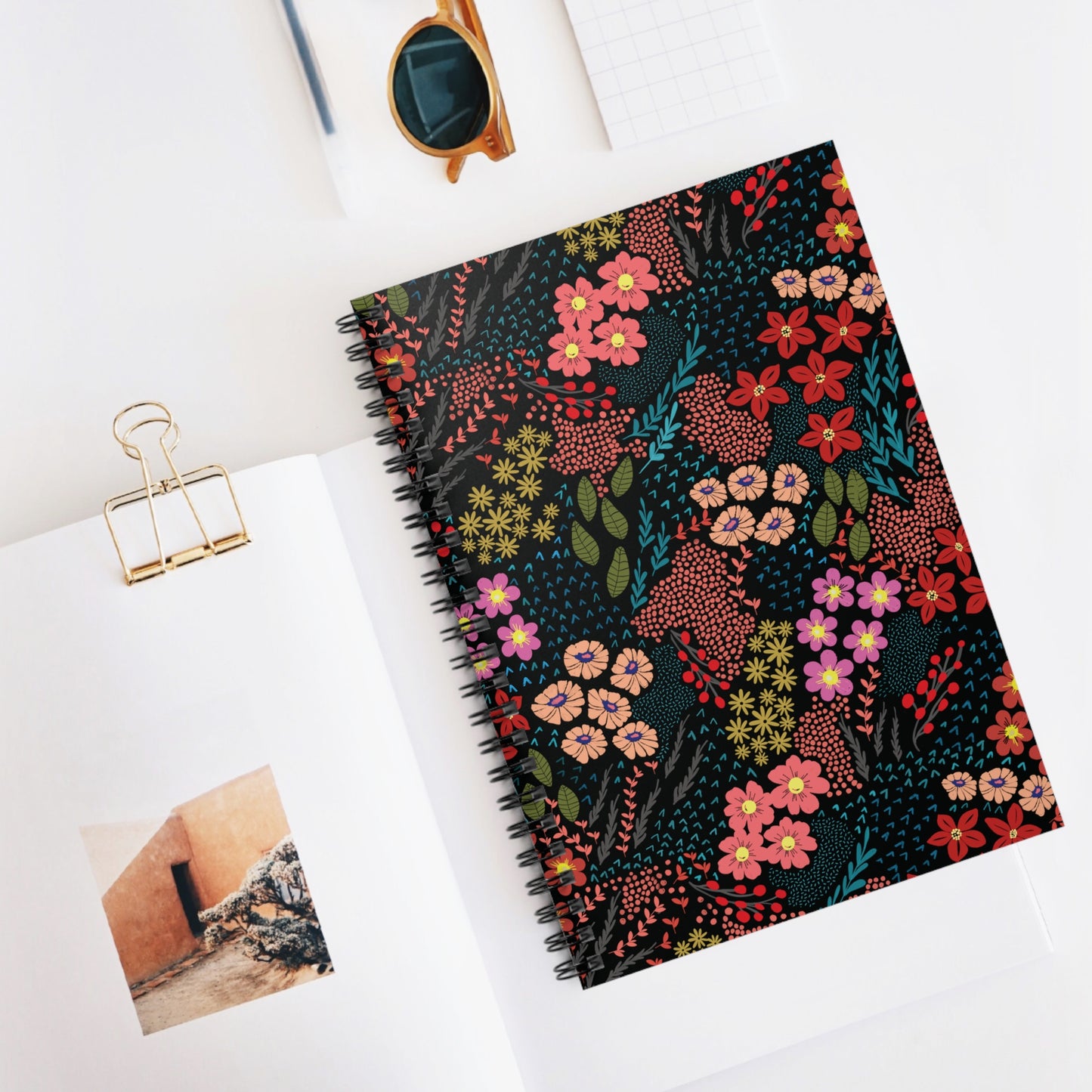Floral Spiral Notebook with vibrant patterns, central on a white desk, atop an open book. Accompanied by sunglasses, a gold binder clip, and a grid-pattern notepad, styled for inspiration.