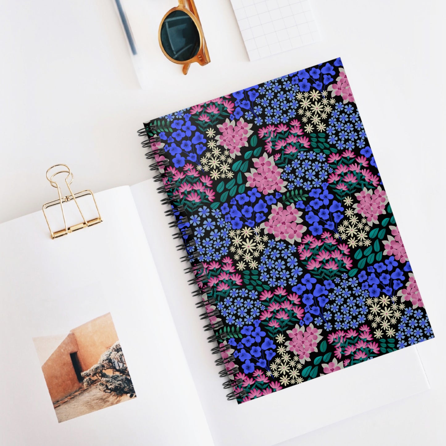 Floral Spiral Notebook with vibrant floral cover, central on a desk styled with a gold-clipped open book, grid notepad, and amber sunglasses, embodying FireBrook Essentials' blend of style and positivity.