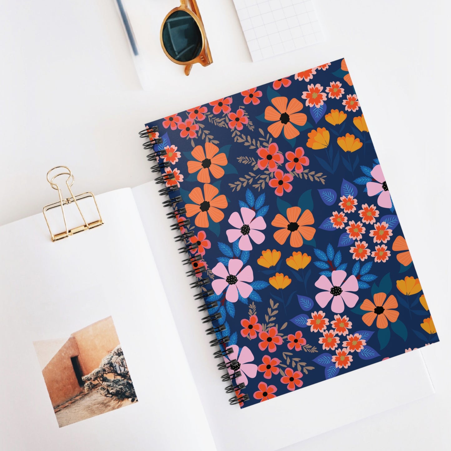 Spiral Notebook, Durable Cover, Ruled, 6x8, featuring bright floral patterns on a dark background, sits centrally in a desktop arrangement with a gold binder clip, round sunglasses, and an open magazine.
