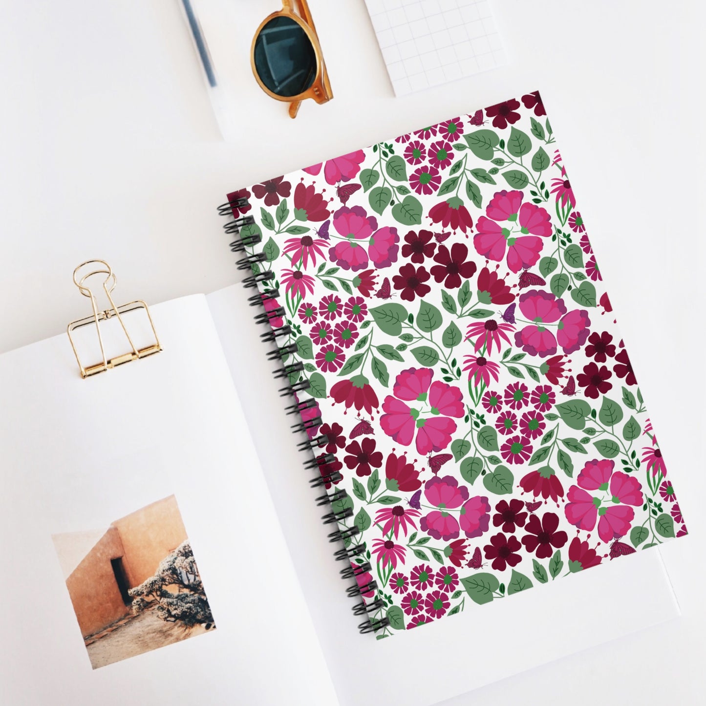 Floral Spiral Notebook elegantly displayed in a flat-lay scene, showcasing its vibrant floral cover and sleek metal binding, perfect for creative note-taking. Nearby, sunglasses and a binder clip complete the stylish setup.