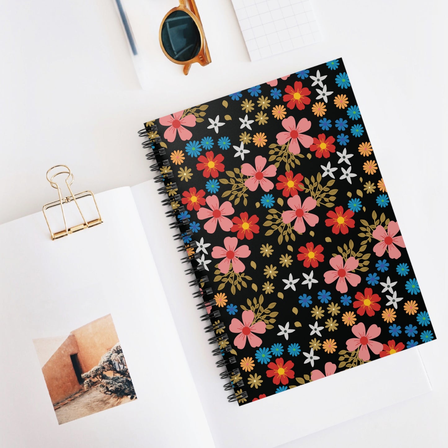 Floral Spiral Notebook with vibrant flowers on the cover, resting atop an open magazine, surrounded by stylish desk items, reflecting FireBrook Essentials' joyful and uplifting design ethos.