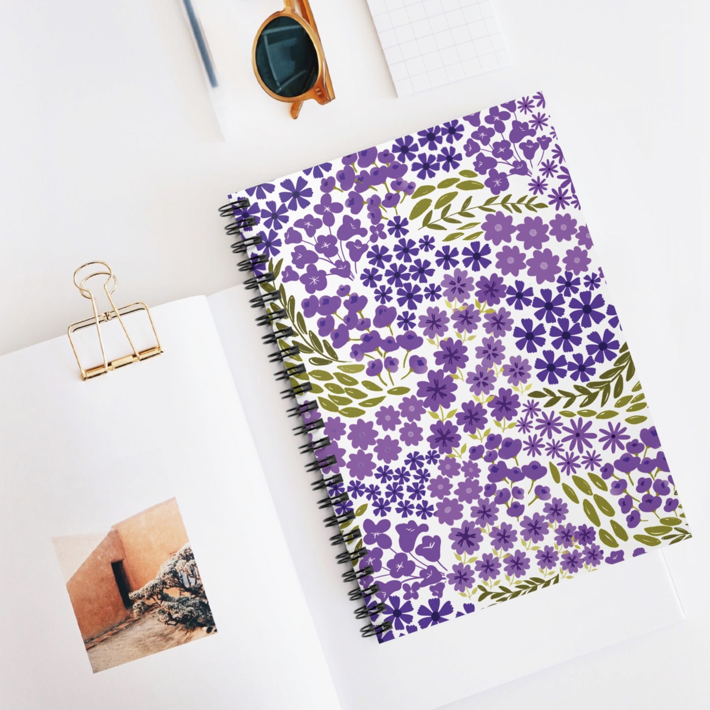 Floral Spiral Notebook with purple flowers and olive leaves on the cover, displayed with sunglasses and an open sketchbook on a white surface, embodying FireBrook Essentials' uplifting and joyful design ethos.