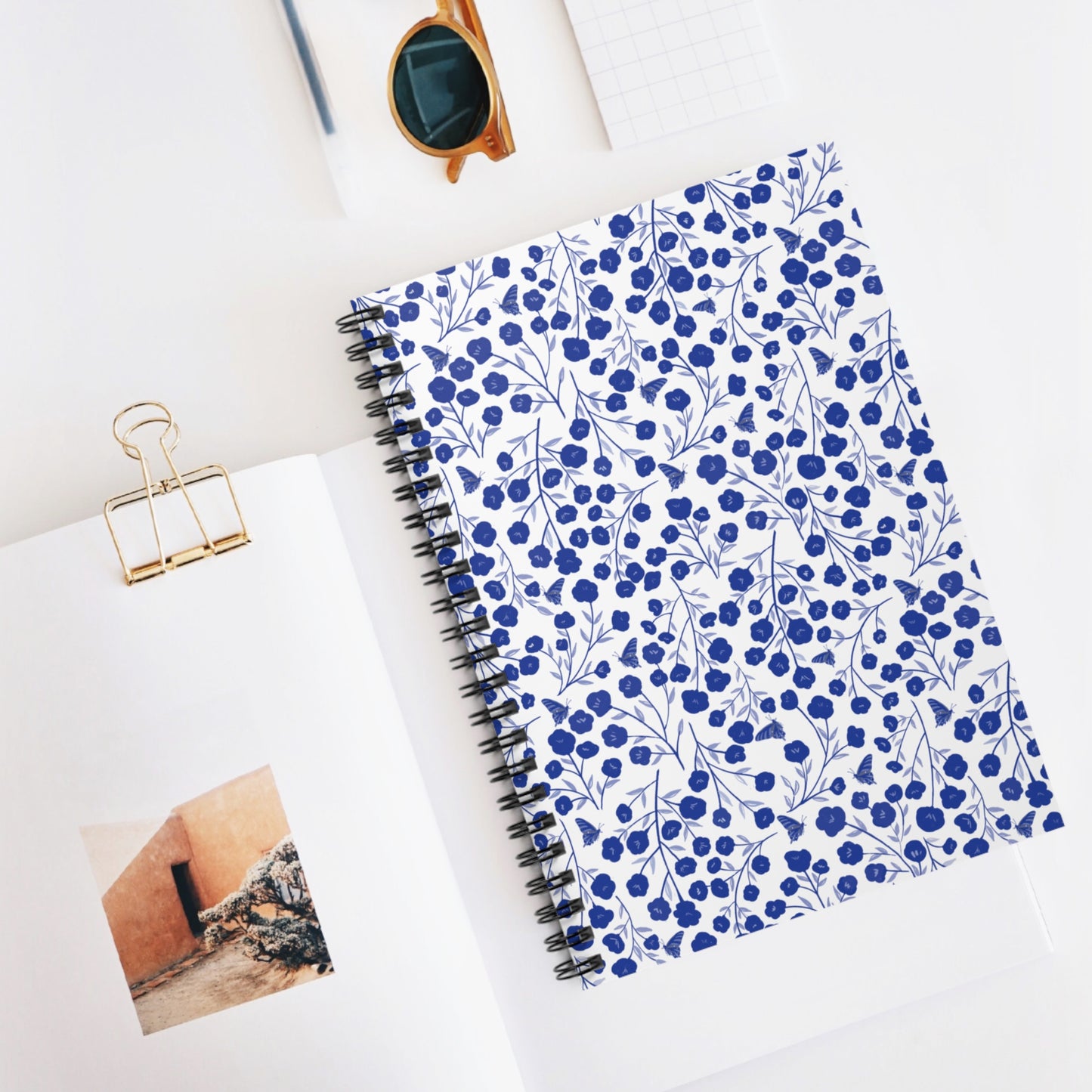 Floral Spiral Notebook with blue flowers and leafy patterns, neatly styled with an open book and sunglasses, epitomizes FireBrook Designs’ blend of style and functionality for creative expression and organization.