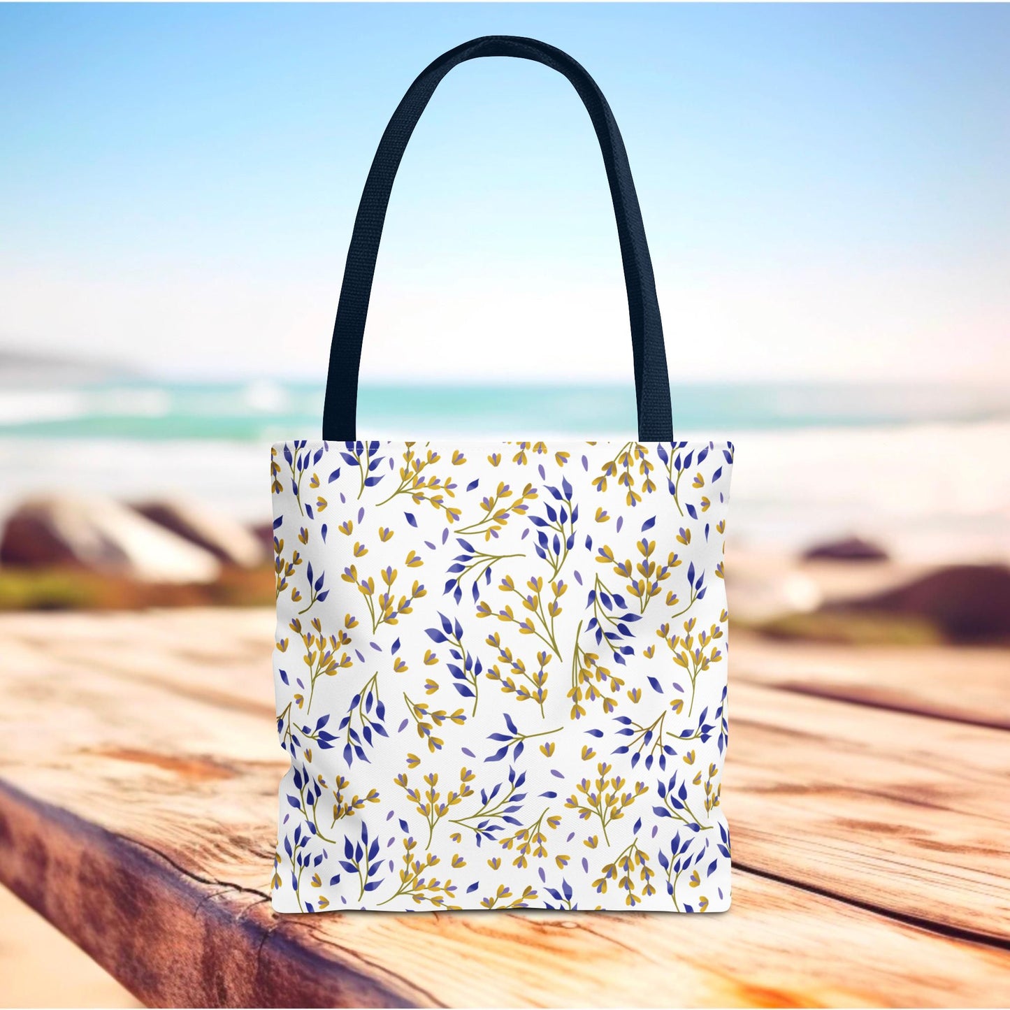 Floral Tote Bag with black handles featuring a gold and royal blue botanical pattern on a white background, resting on wood with a beach backdrop, ideal for joyful summer outings.
