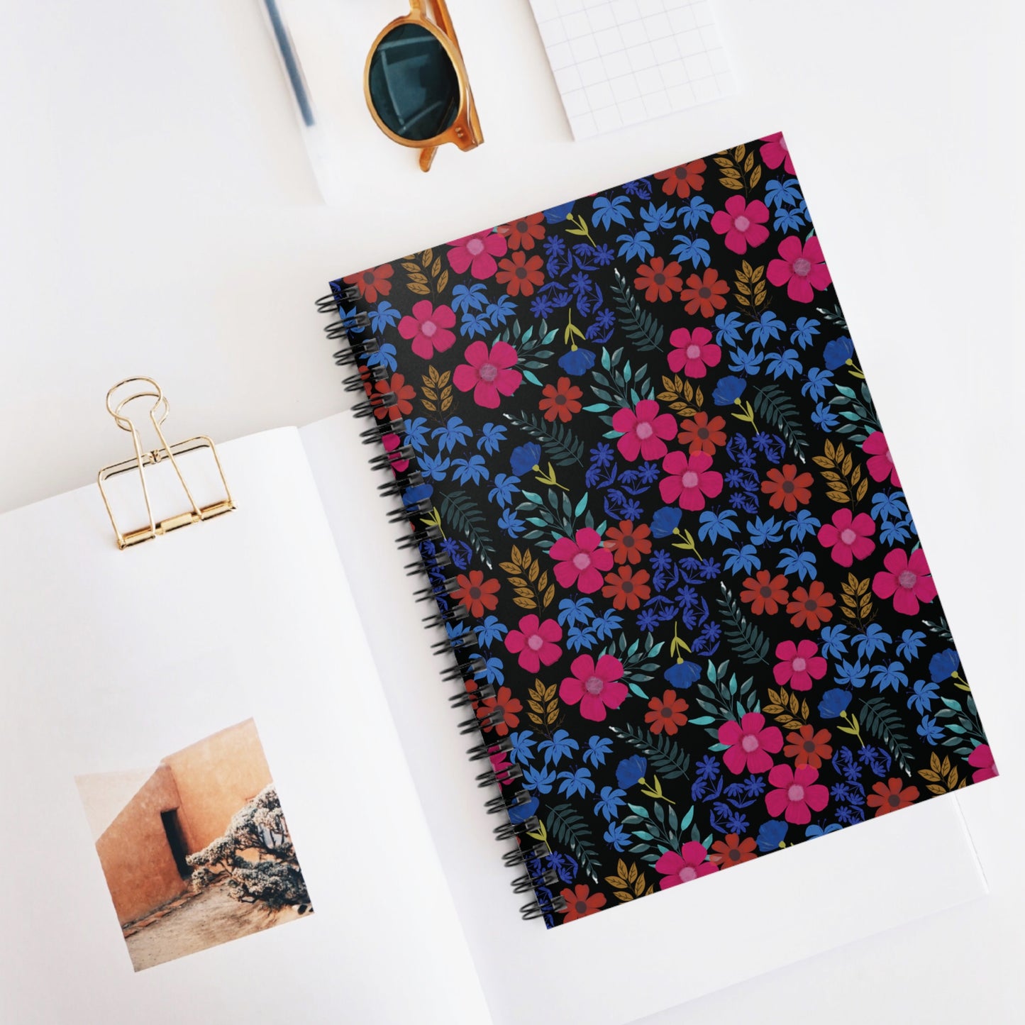 Floral Spiral Notebook with vibrant flower design, round sunglasses, and a grid notepad on a white workspace; embodies FireBrook Designs' joyful, uplifting style for creative note-taking.