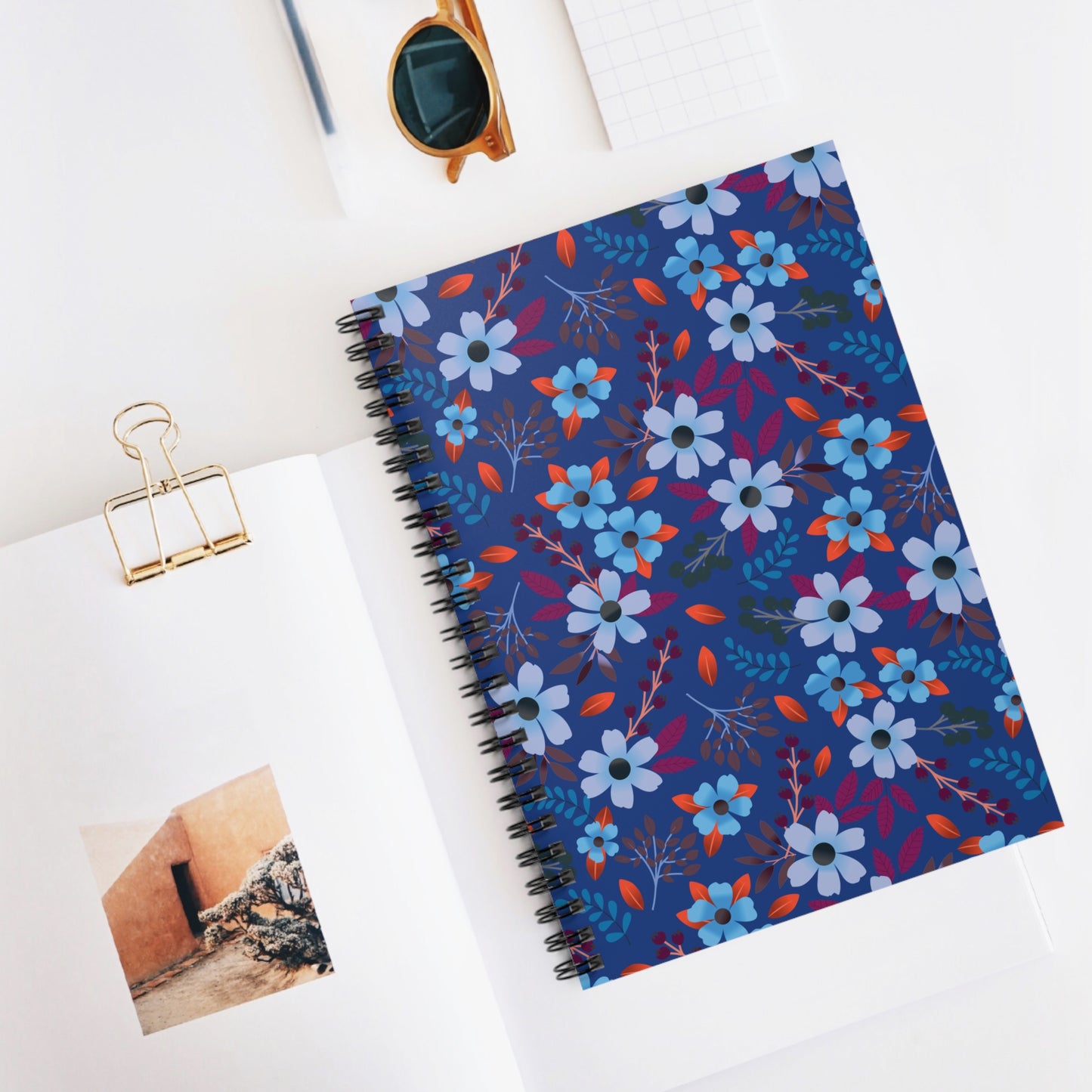 Floral Spiral Notebook with bold blue flowers and red-orange leaves on a navy cover, centered on a tidy white desktop, showcasing FireBrook Designs' uplifting and creative style.