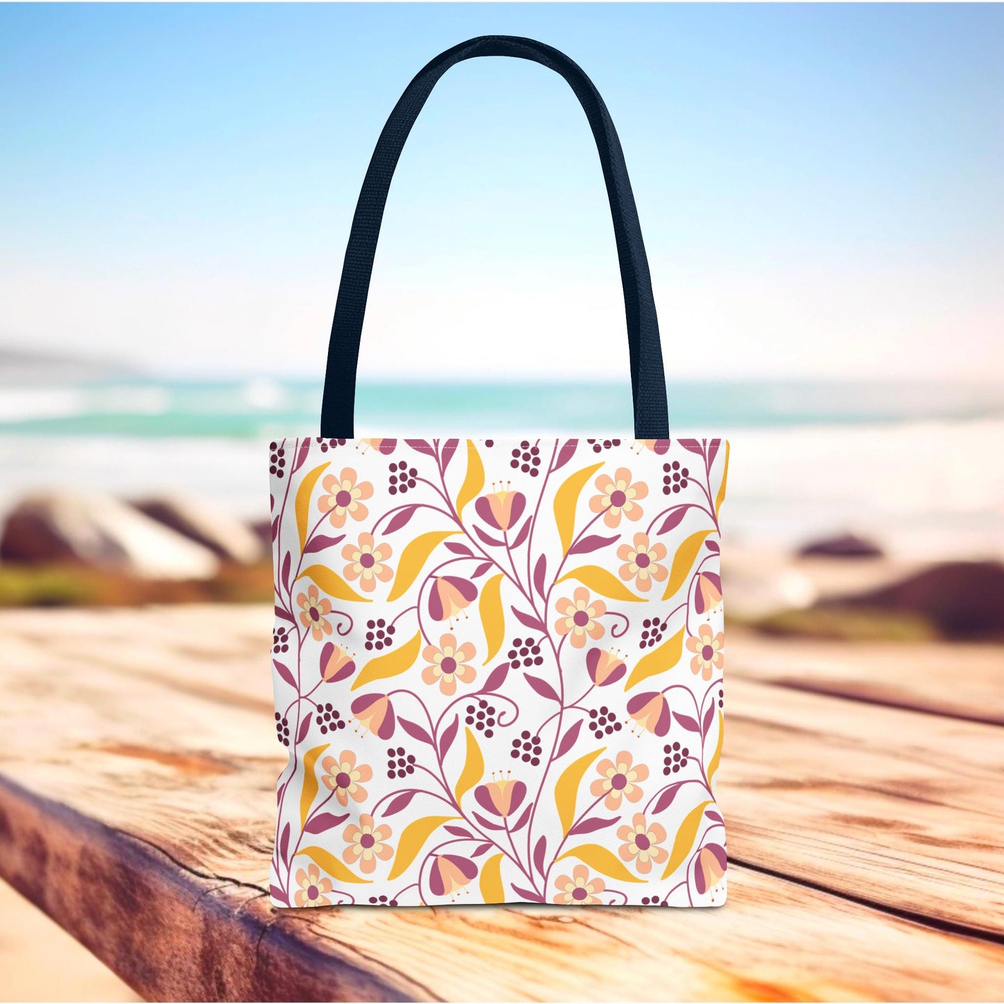 Floral Tote Bag with dark straps features a cheerful floral motif in pinks, mustard yellow, and burgundy, ideal for summer outings. Displayed on a wooden surface with ocean background.