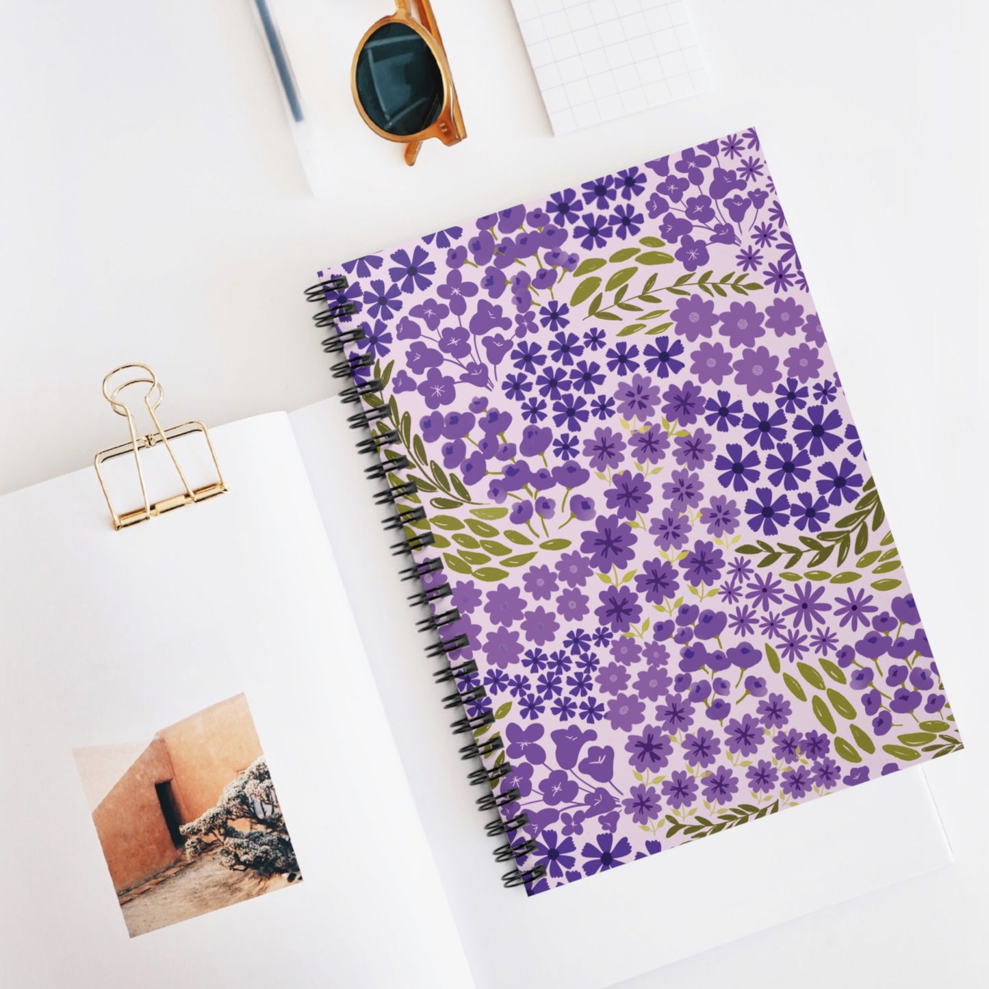 Floral Spiral Notebook with purple flowers and olive leaves on a pink cover, surrounded by an open book, gold binder clip, amber sunglasses, and a grid-patterned notepad corner, embodying FireBrook Designs' joyful style.