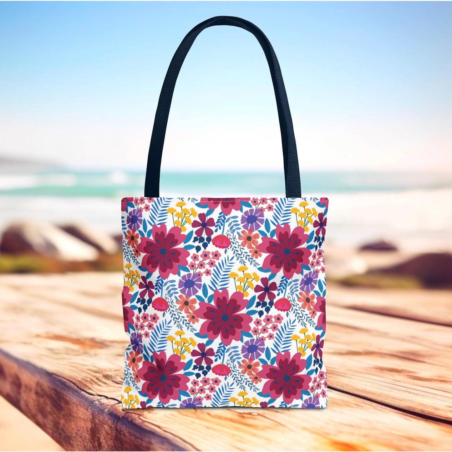 Floral Tote Bag with vibrant magenta, purple, and yellow blooms on a white background, resting on a wooden surface. Features sturdy dark handles, ideal for summer outings or beach trips.