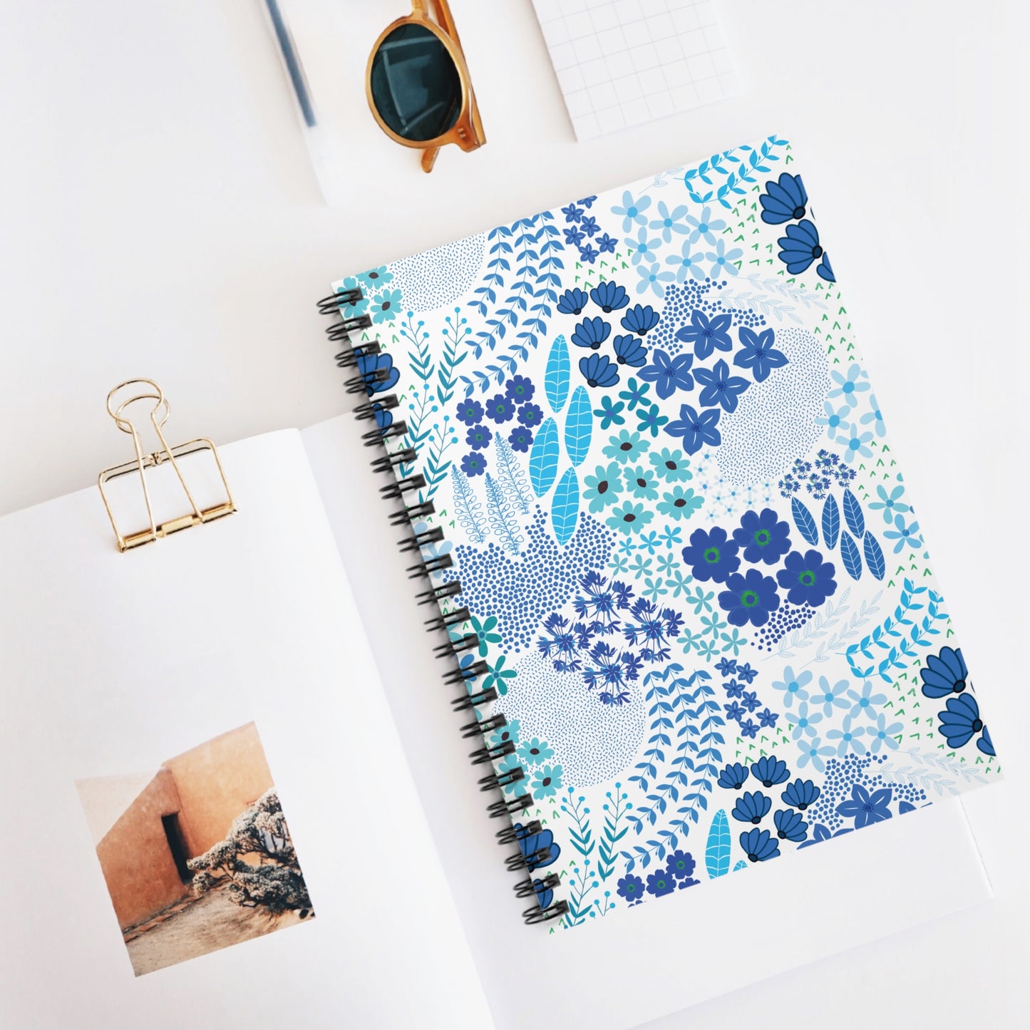 Floral Blue Motif Spiral Notebook with vibrant floral cover, centered on a white workspace with sunglasses and an open book nearby, embodying FireBrook Designs' uplifting, creative spirit. Ideal for notes and sketches.
