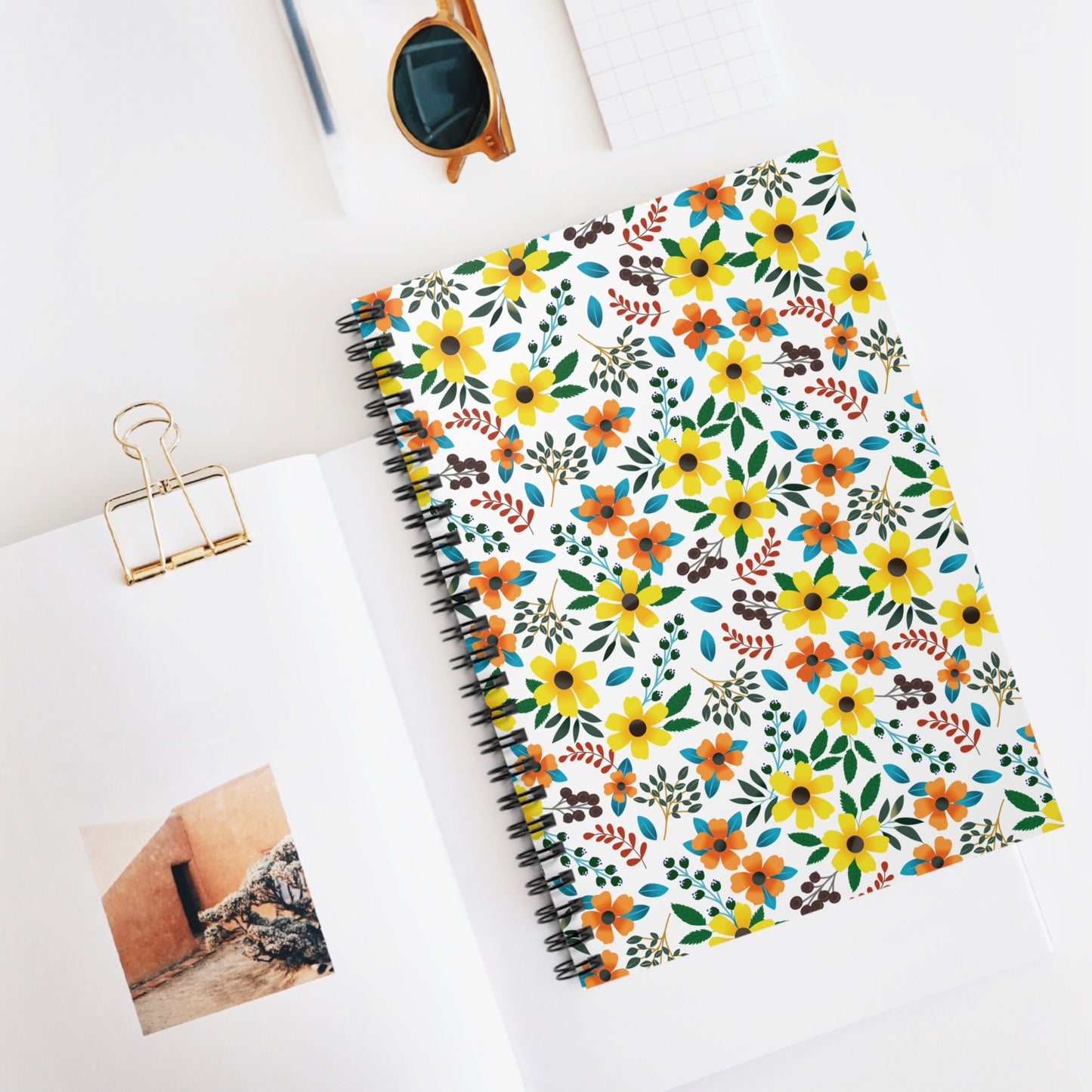 Floral Spiral Notebook featuring yellow and orange flowers with green leaves on its cover, set in a tidy, bright workspace alongside an open book and tortoiseshell sunglasses, embodying FireBrook's joyful, uplifting designs.