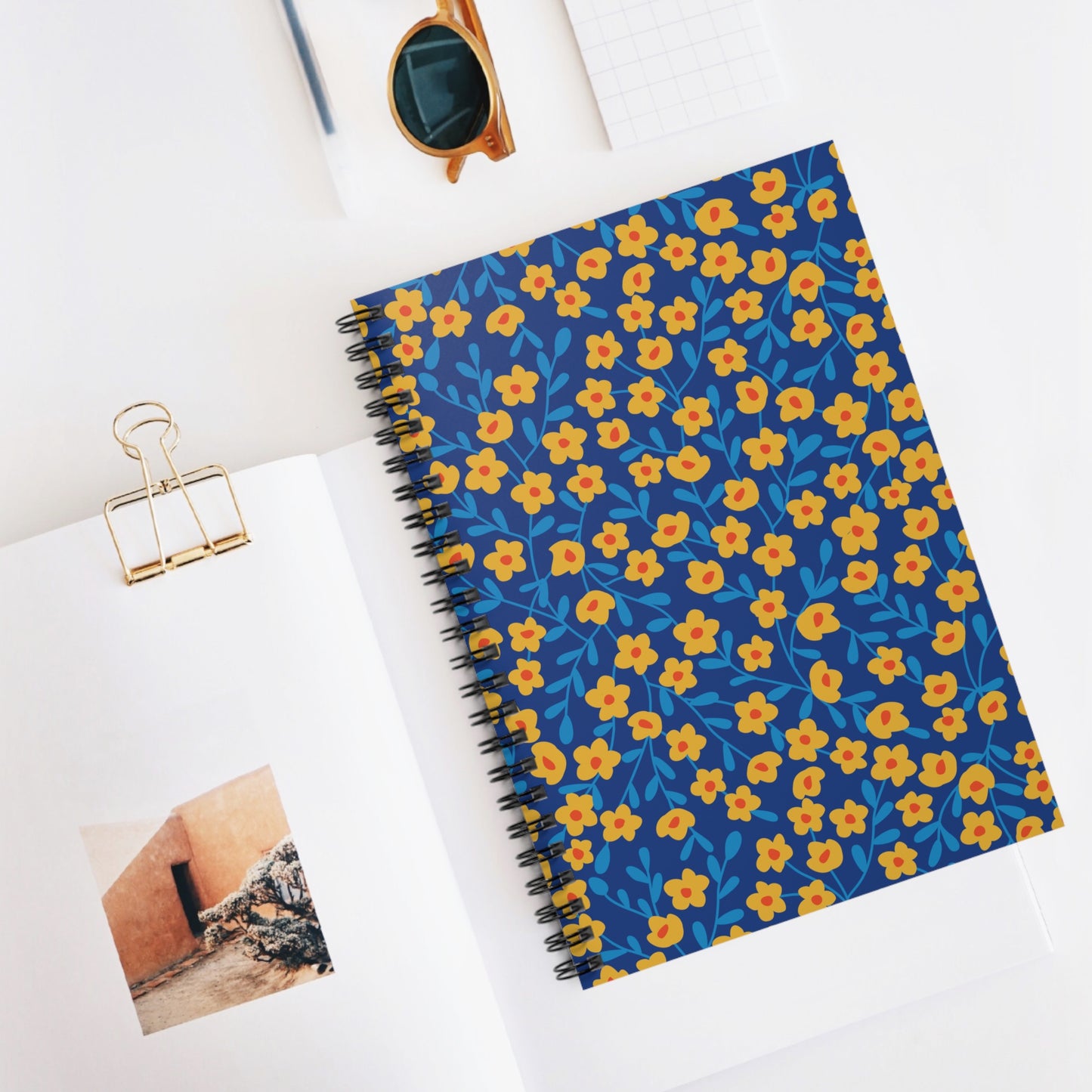 Spiral Notebook, Durable Cover, Ruled, 6x8 – Sunlit Blossoms (Blue Background), featuring a charming floral pattern, lies on a workspace beside a gold binder clip, sunglasses, and open reading material.