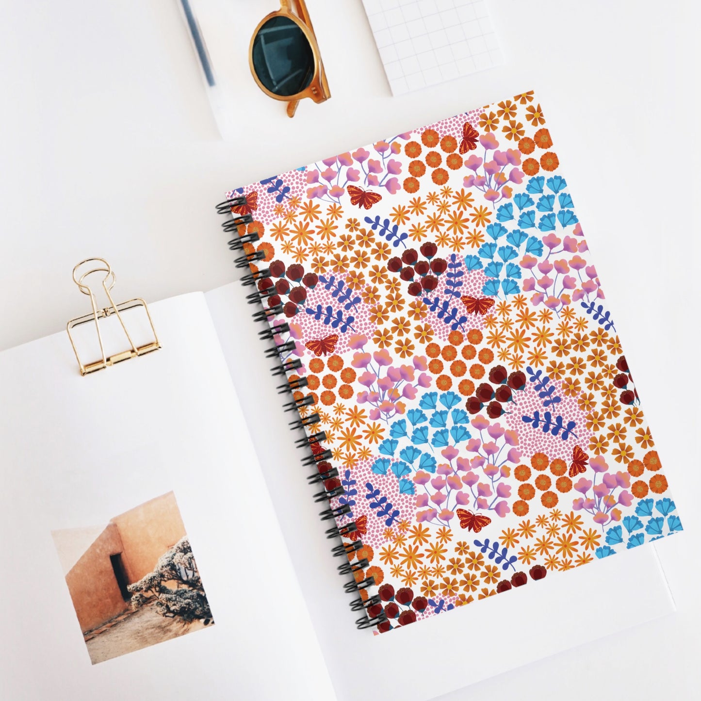 Floral Spiral Notebook with vibrant floral and butterfly cover, sits on a white desk beside an open magazine and sunglasses. Ideal for creative tasks, reflecting FireBrook Essentials' joyful and uplifting design ethos.