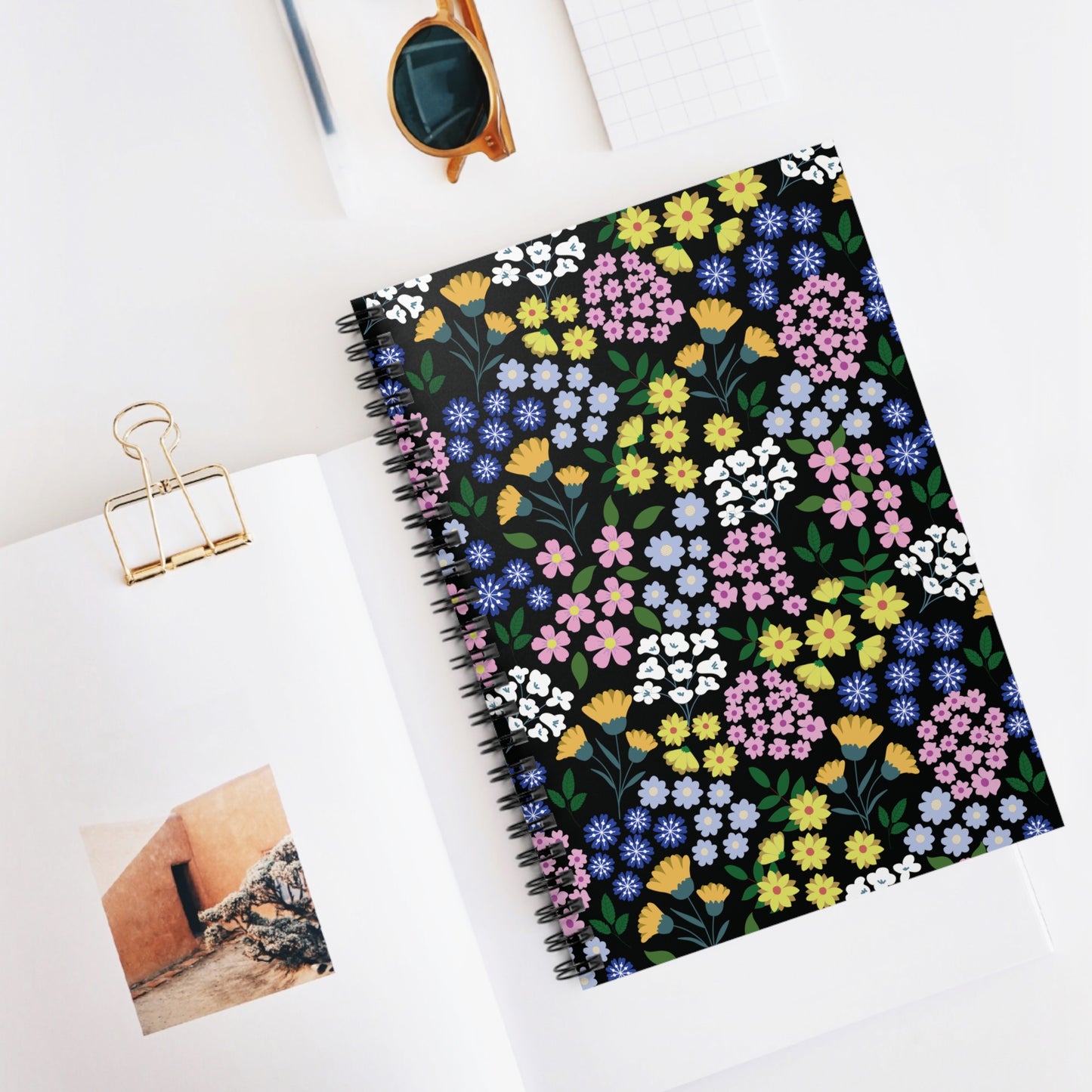 Spiral Notebook, Durable Cover, Rules, 6x8, featuring a flower-patterned cover on a tidy workspace; surrounded by an open book, amber-framed sunglasses, and a grid-page notepad, embodying FireBrook Essentials' joyful design ethos.