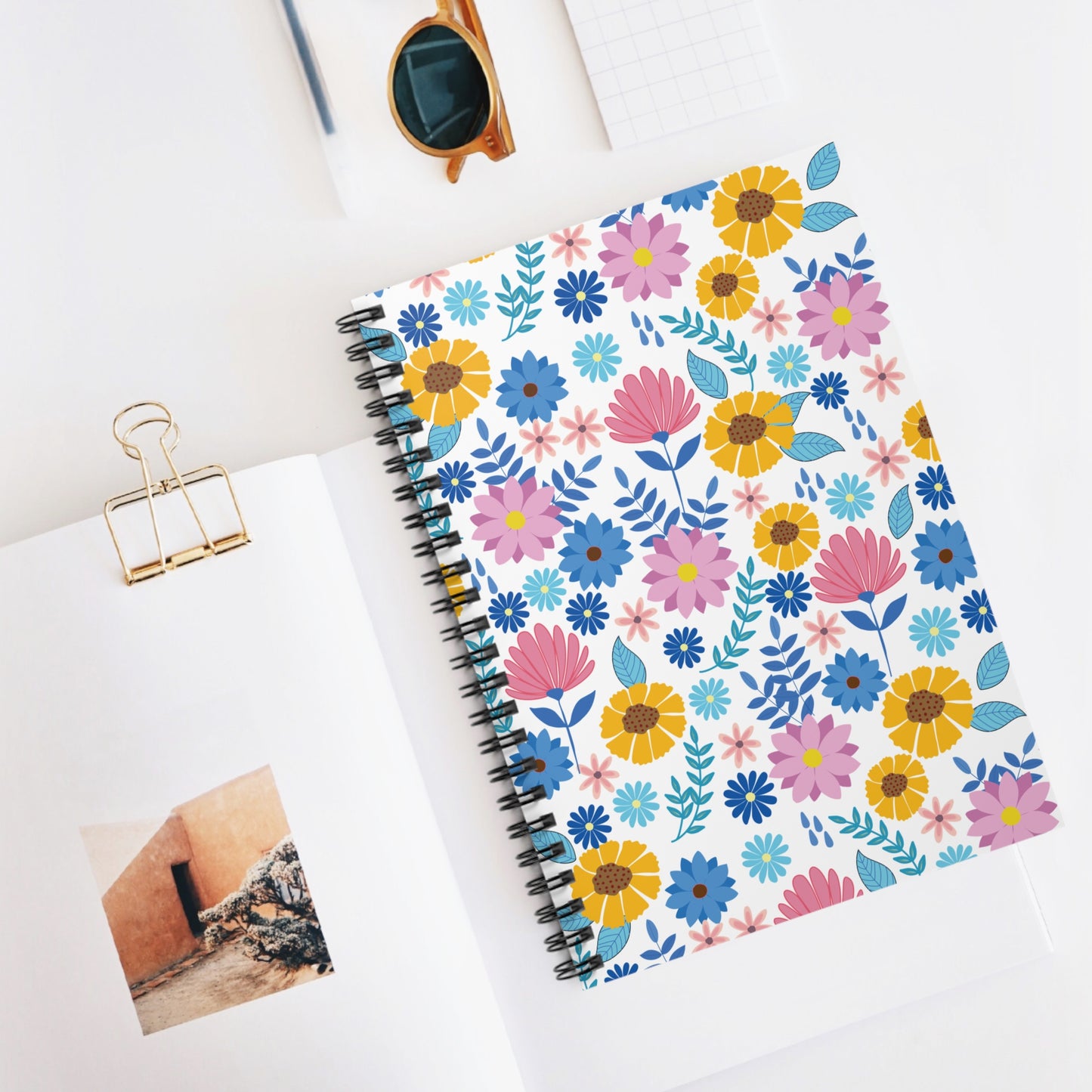 SPIRAL NOTEBOOK, DURABLE COVER, RULED, 6X8 featuring a vibrant floral design, centered on a white desktop beside a gold binder clip, sunglasses, and a small notepad, embodying FireBrook Essentials' joyful style.