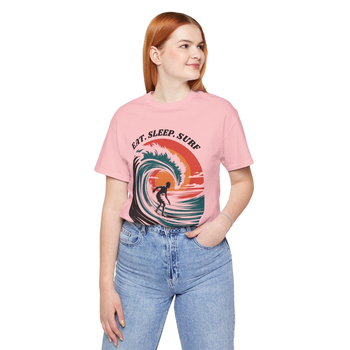 Woman in a pink Surf Tee - Eat Sleep Surf Design with a surfer print, paired with blue jeans. The tee offers a casual, beachy vibe, perfect for surf enthusiasts.