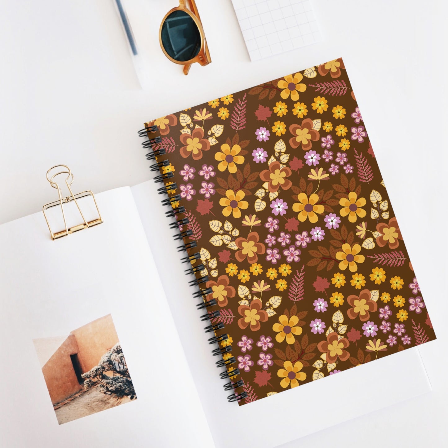 SPIRAL NOTEBOOK, DURABLE COVER, RULES, 6X8 - RETRO BLOOM, BROWN BACKGROUND, featuring warm-toned floral design. Positioned on an open publication, with nearby sunglasses and a grid-patterned notepad, reflecting FireBrook Designs' joyful style.