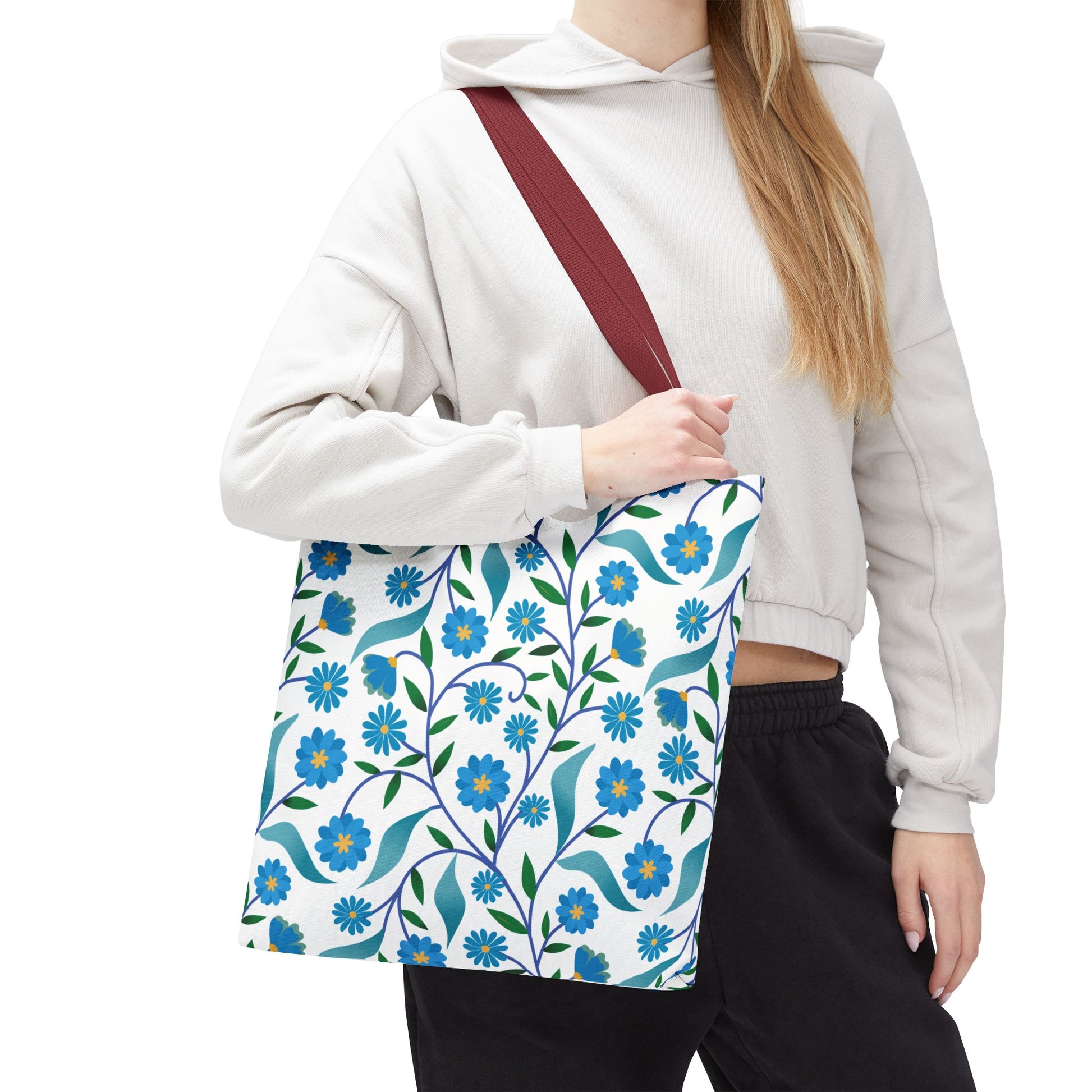 Person wearing a light-colored cropped hoodie and dark pants carries a Blue Floral Tote Bag, featuring a white background with blue flowers and green leaves, over their shoulder with a dark-red strap.