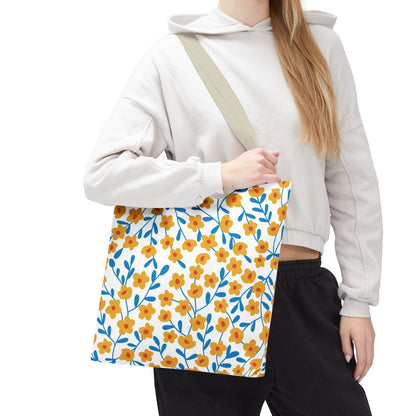 Buttercup Cream Tote Bag with small yellow-orange flowers and blue leaves, held by a person in casual attire. Ideal for joyful summer outings, featuring durable polyester, reinforced handles, and spacious boxed corners.