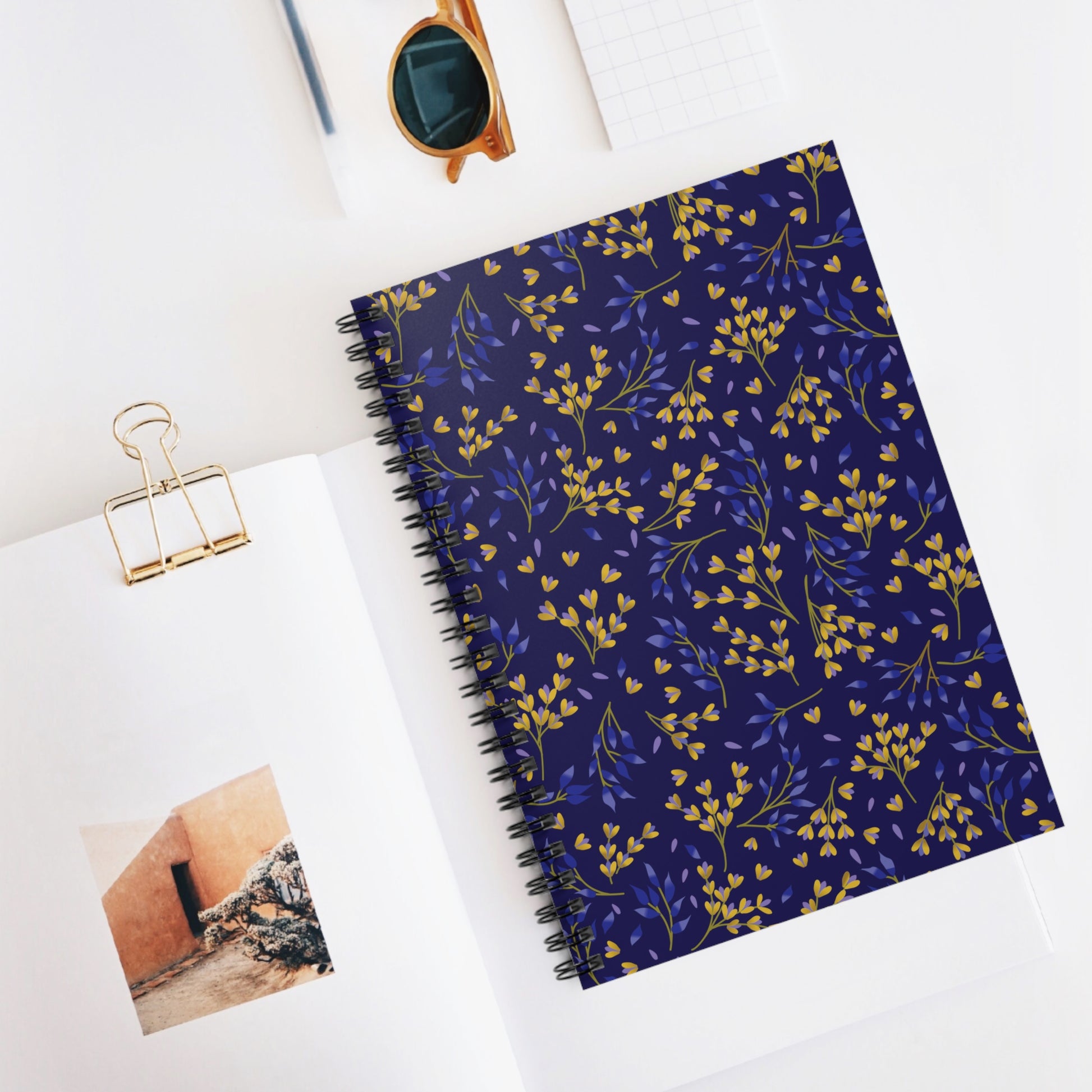 Floral Spiral Notebook with navy cover adorned with yellow and blue floral sprigs, ideal for creativity and organization. Features sleek metal spiral binding, shown with a magazine and amber sunglasses for a stylish touch.
