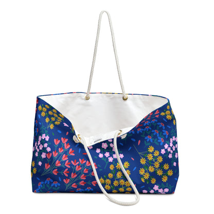 Weekender Bag featuring a deep blue floral design with red, pink, and yellow blossoms. It has white rope handles through gold-tone grommets and a T-bottom for added space. Ideal for stylish travel.