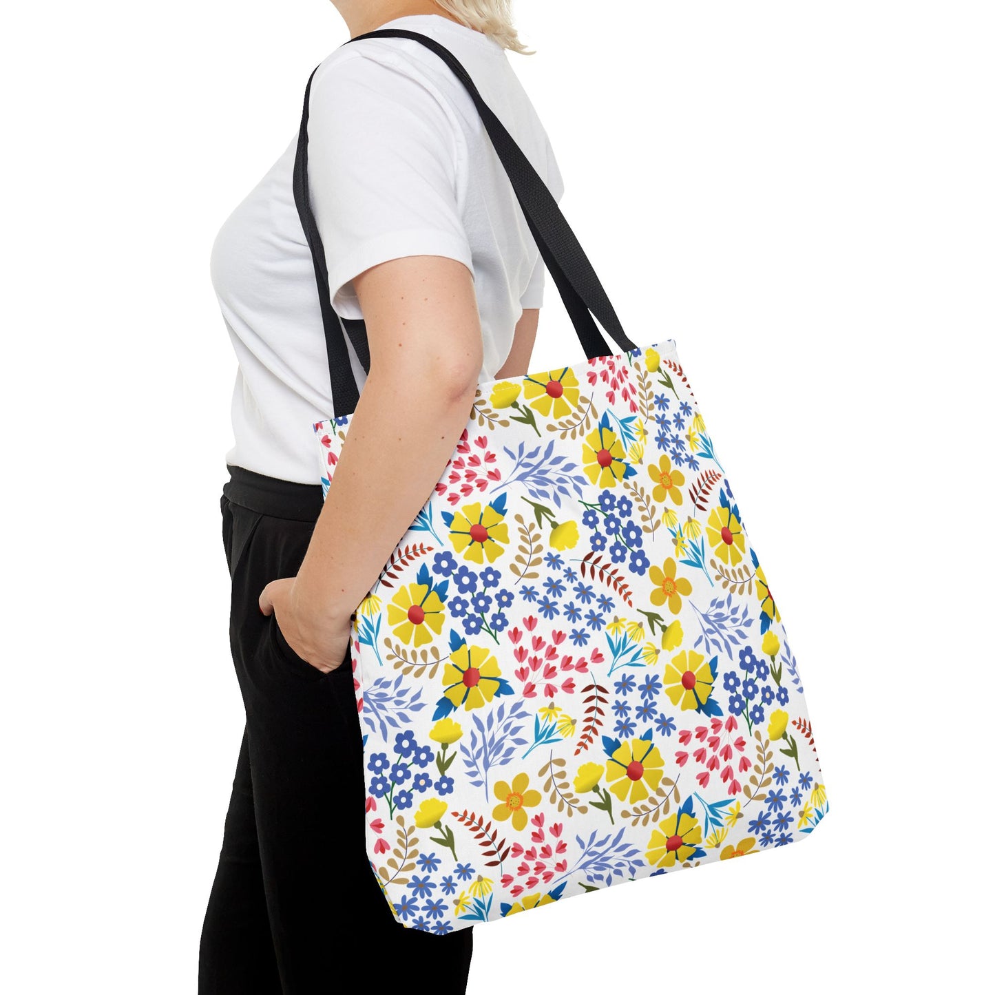 A person carries a Floral Tote Bag from FireBrook Designs, featuring a vibrant multicolored floral pattern on a white polyester body, complemented by black straps, perfect for joyful summer outings.