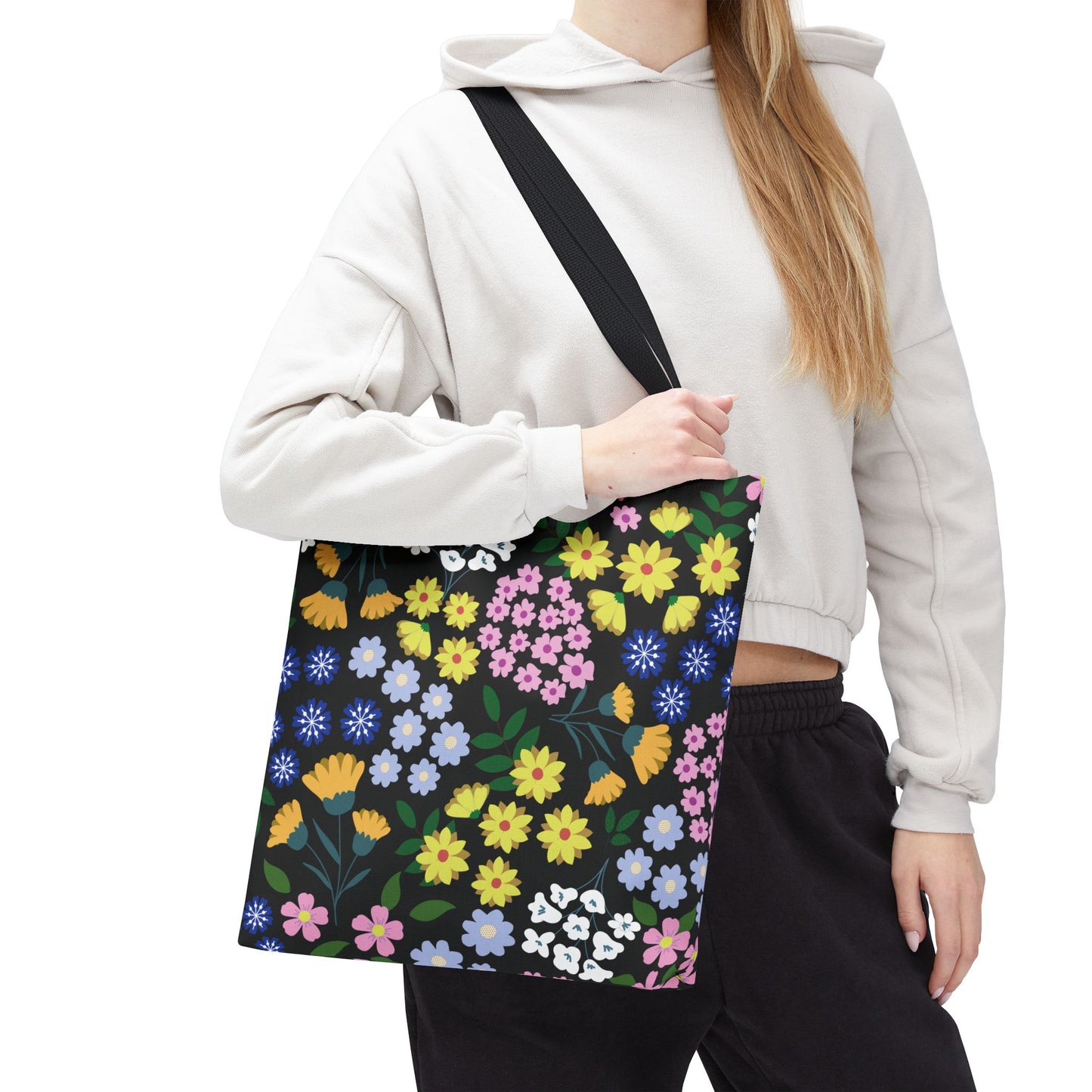 Person wearing a cropped hoodie and dark pants, holding the Starry Night Blooms Tote Bag featuring a vibrant floral pattern with daisies and blossoms, showcasing its cheerful design and durable, spacious build.