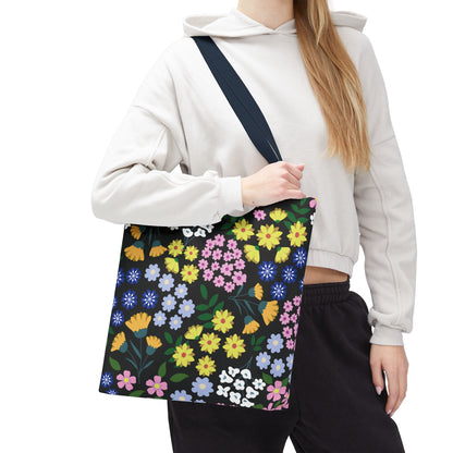 Person carrying the Starry Night Blooms Tote Bag, a black canvas tote with a vibrant floral print of yellow, pink, blue, and white flowers, ideal for cheerful summer outings.