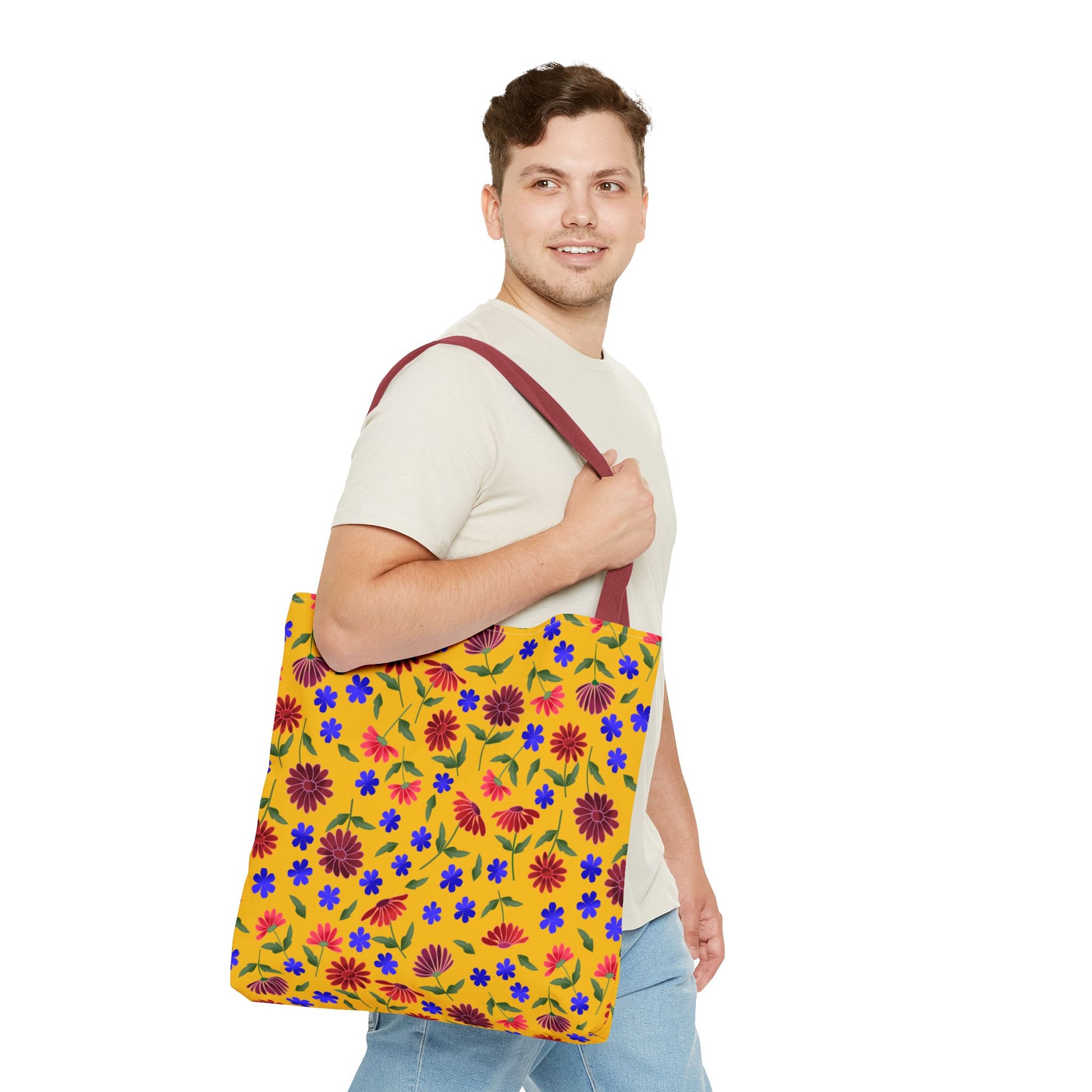 A young man models the Golden Wildflower Tote Bag, featuring vibrant red, purple, and blue floral patterns on a yellow background, complete with sturdy red straps, embodying FireBrook Designs’ joyful, practical style.