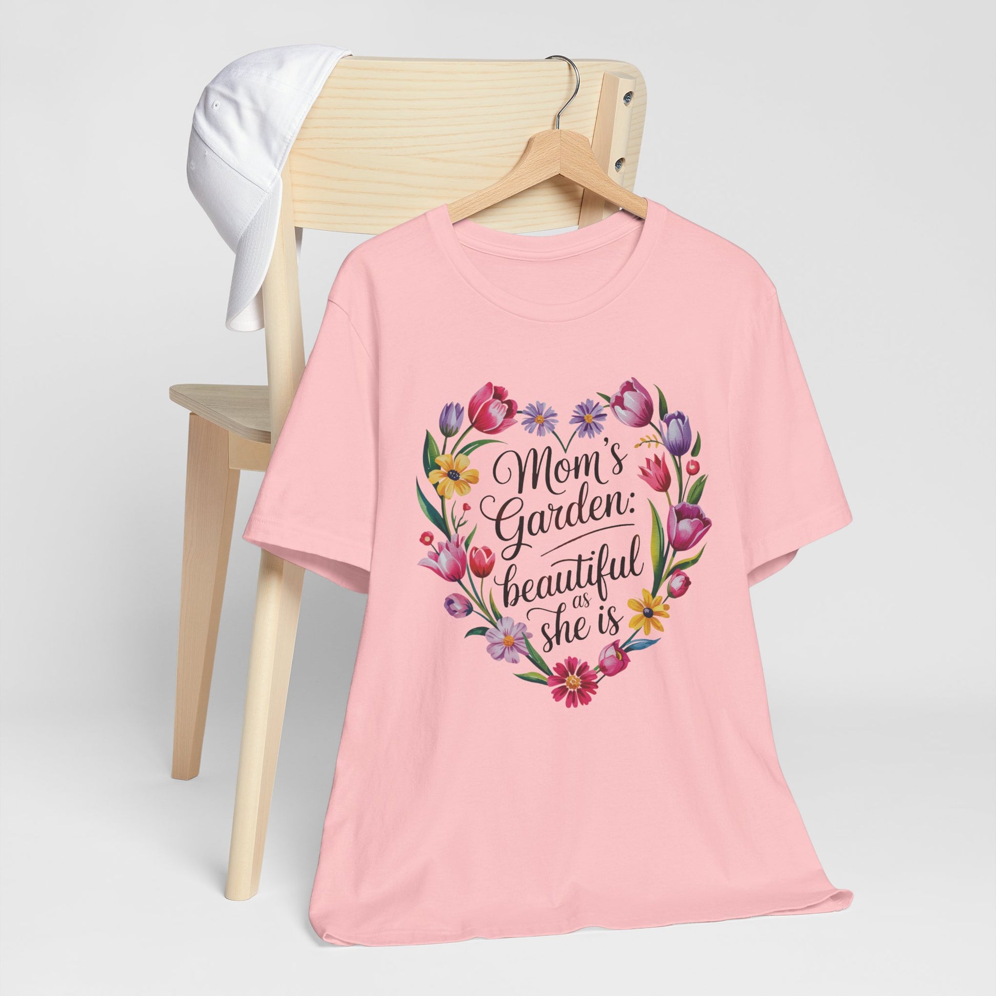 Mom's Garden is as Beautiful as She Is Mother's Day Comfort Colors Unisex T-Shirt, featuring a heart-shaped floral design, draped over a chair with a white hat.