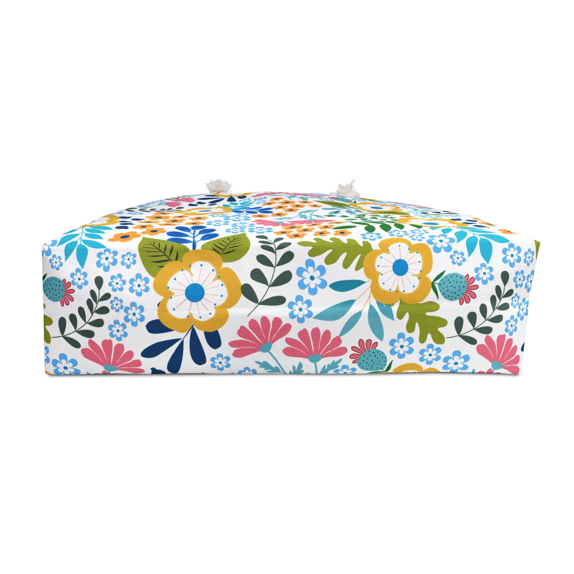 Weekender Bag displayed as a small upholstered floor pouf with a vibrant floral print of yellow, pink, and blue flowers on white fabric, featuring tassel-style knots on top corners.