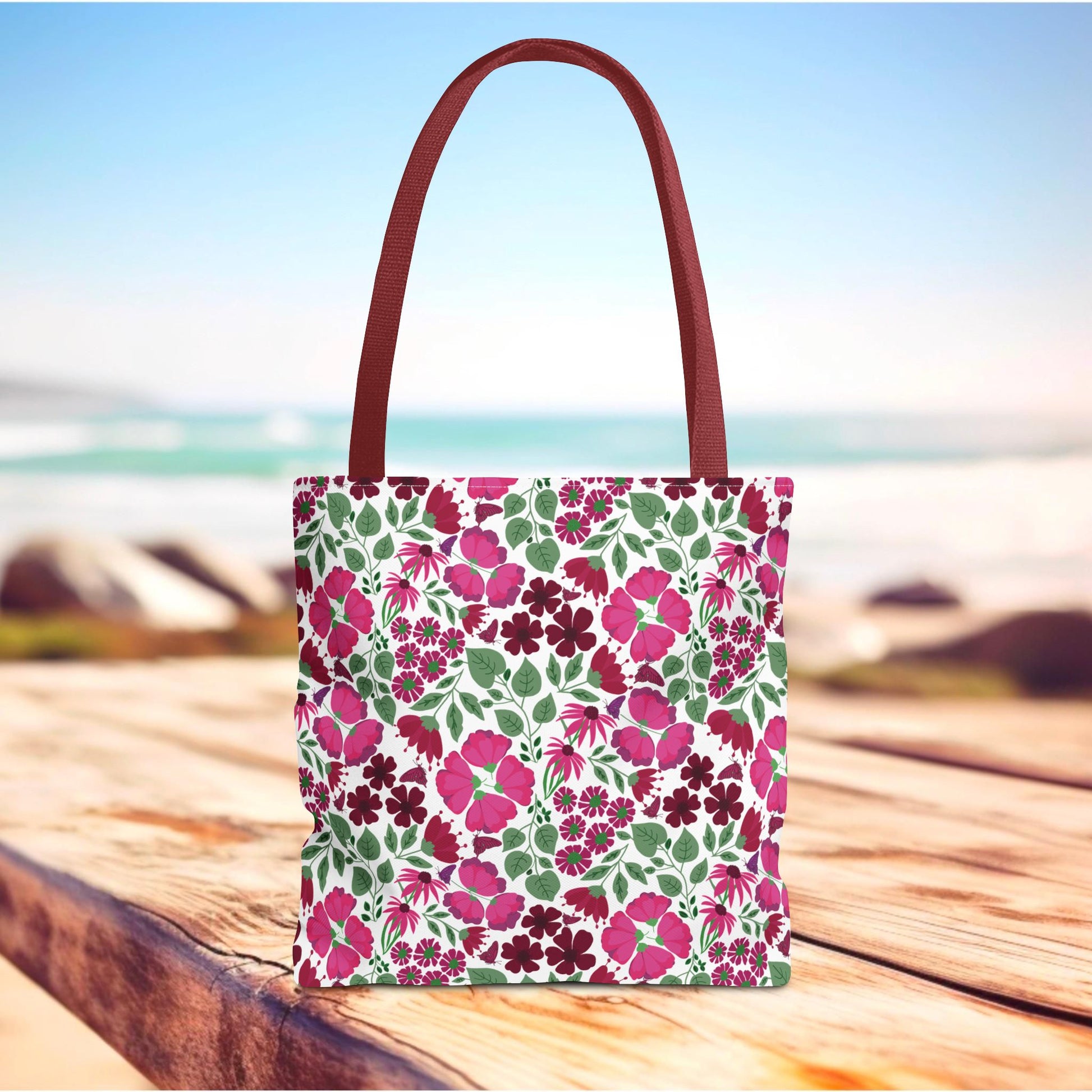 Pink Lady Tote Bag with a vibrant pink and berry floral design, featuring sturdy burgundy handles, standing beautifully against a soft beach backdrop. Ideal for adding cheerful style to daily outings.