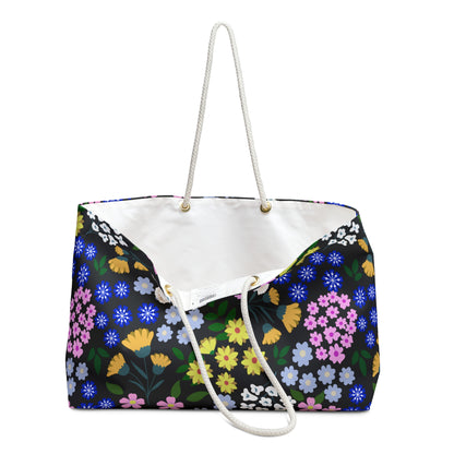 Weekender Bag with multicolor floral pattern on black, featuring white braided rope handles through gold grommets. Spacious, stylish design with cream interior, perfect for joyful travels.