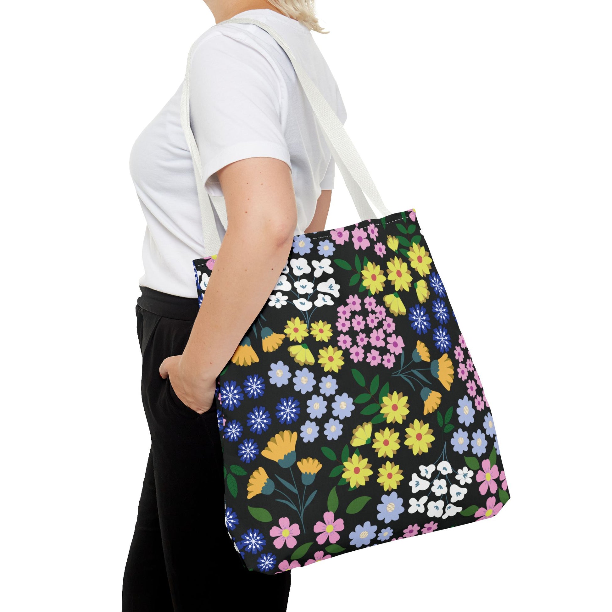 Floral Tote Bag with vibrant blossoms in yellow, pink, blue, and purple on black, carried by a person in casual attire. Ideal for summer, it combines style and practicality from FireBrook Designs.
