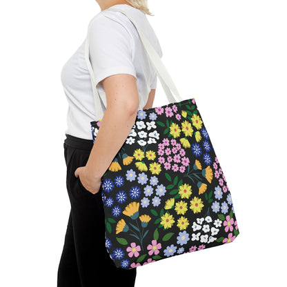 Floral Tote Bag with vibrant blossoms in yellow, pink, blue, and purple on black, carried by a person in casual attire. Ideal for summer, it combines style and practicality from FireBrook Designs.
