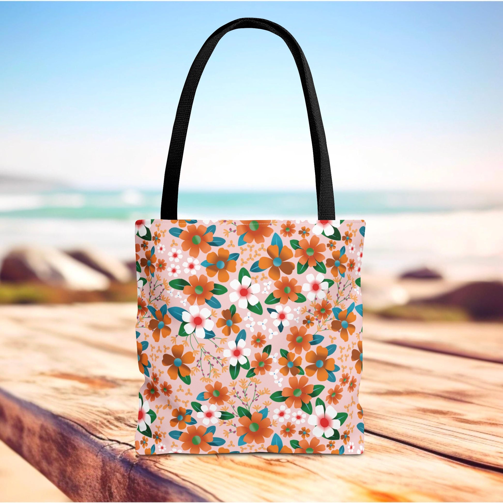 Floral Tote Bag by FireBrook Essentials on a deck, featuring vibrant orange and white blossoms against a peachy-pink background, black handles, ideal for summer outings or beach trips.