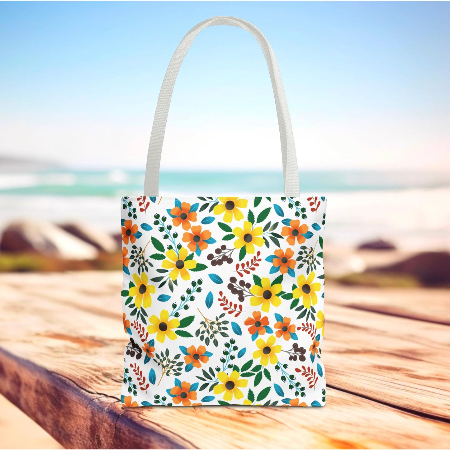 Floral Tote Bag with vibrant yellow and orange flowers, green leaves, and berry-like sprigs on a white background, set on a wooden surface with an ocean backdrop, embodying FireBrook Designs' joyful, stylish essence.