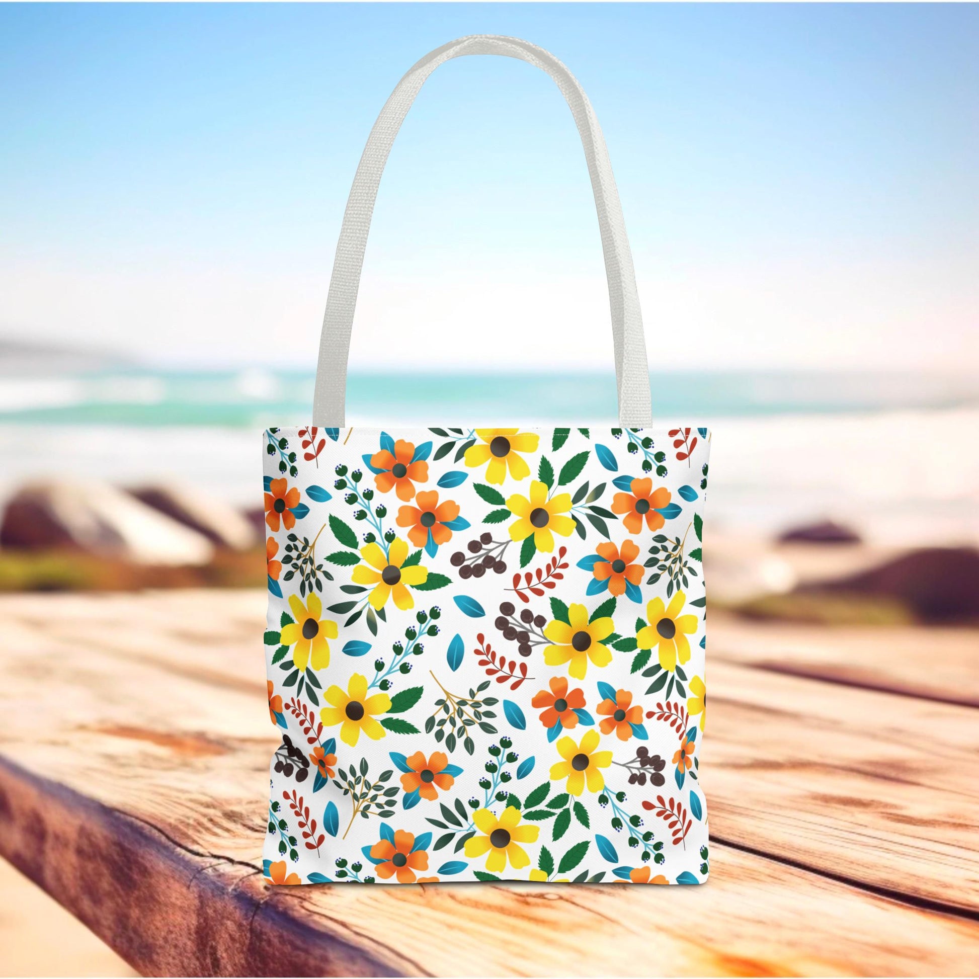 Floral Tote Bag with vibrant yellow and orange flowers, green leaves, and berry-like sprigs on a white background, set on a wooden surface with an ocean backdrop, embodying FireBrook Designs' joyful, stylish essence.