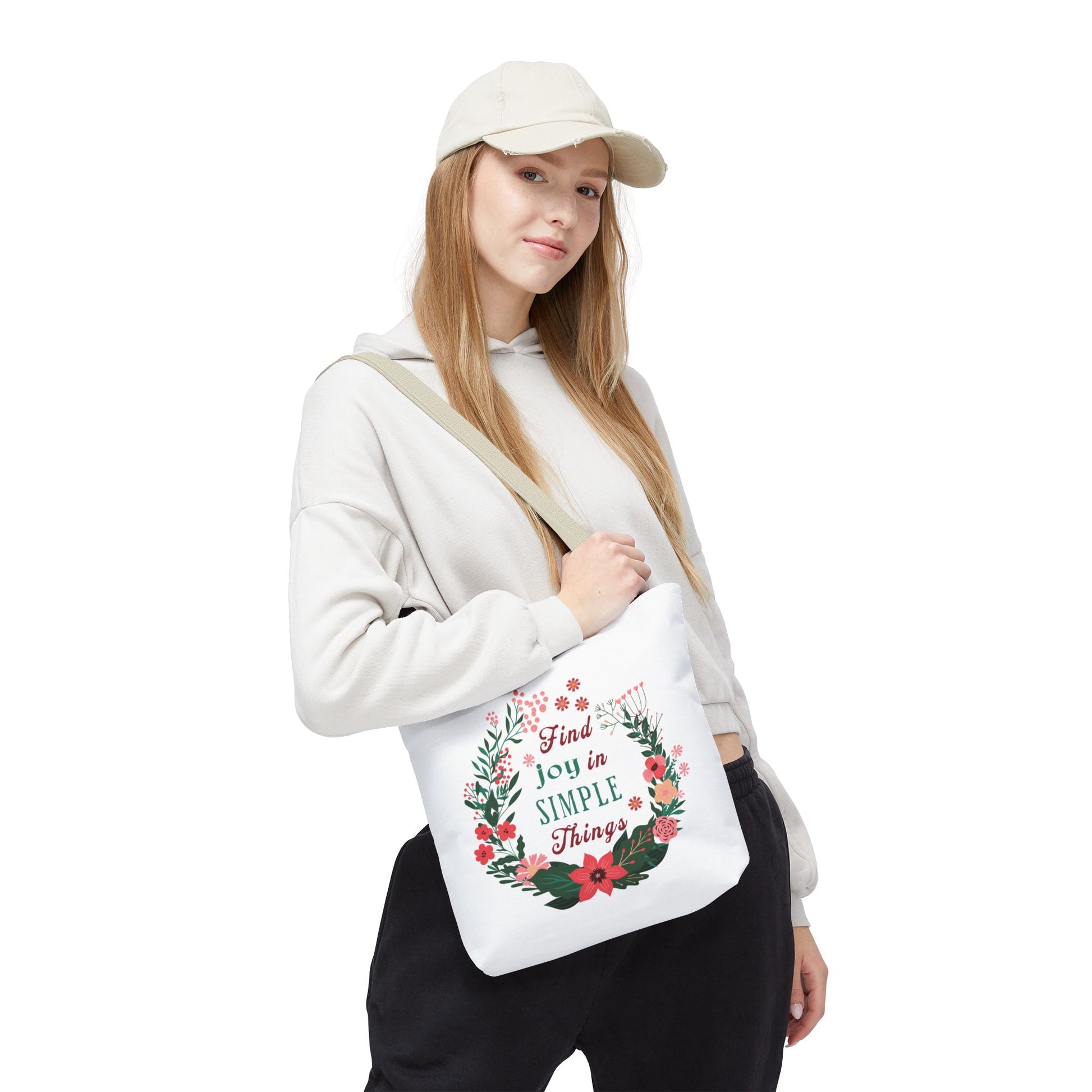 Woman wearing a hat and sweatshirt holding the Find Joy in Simple Things tote bag with a floral design, showcasing its stylish and functional appeal.