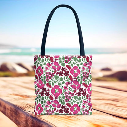 Pink Lady Tote Bag with vibrant pink and berry floral pattern on a white background, featuring black shoulder straps, sits on a wooden surface against a blurred beach backdrop, embodying FireBrook Designs' cheerful, uplifting style.