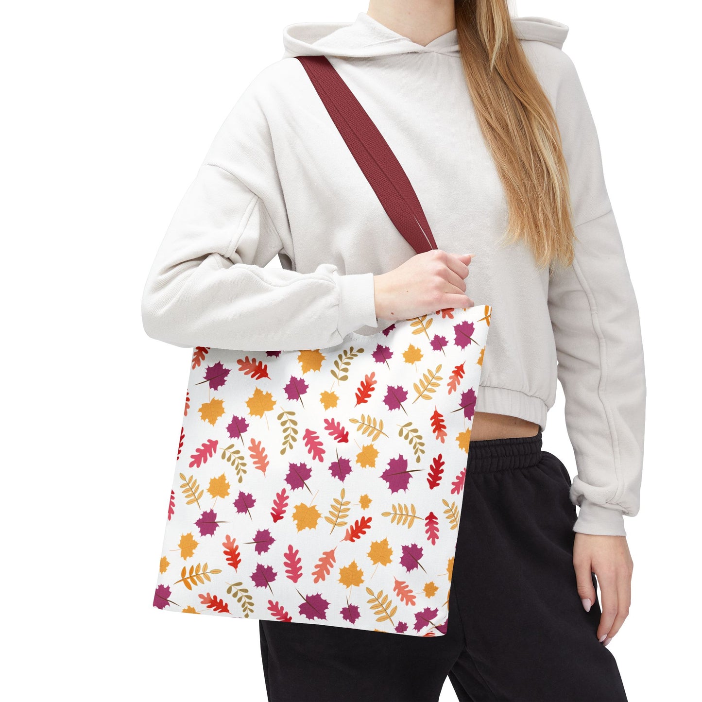 Autumn Leaves Tote Bag with a burgundy strap, featuring a vibrant fall leaf pattern in orange, red, gold, and plum, embraced casually by a person in a light hoodie and dark pants.