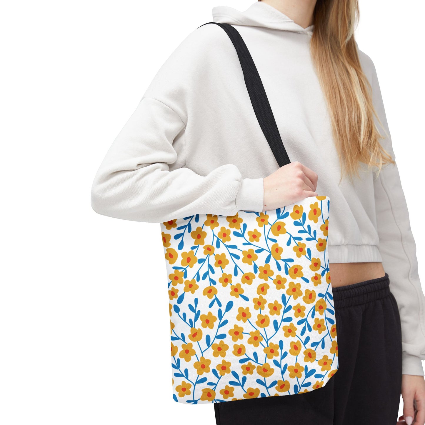 Floral Tote Bag featuring a vibrant pattern of yellow flowers with orange centers and blue stems on a white canvas, carried by a person in a light hoodie. Perfect cheerful accessory for summer outings.