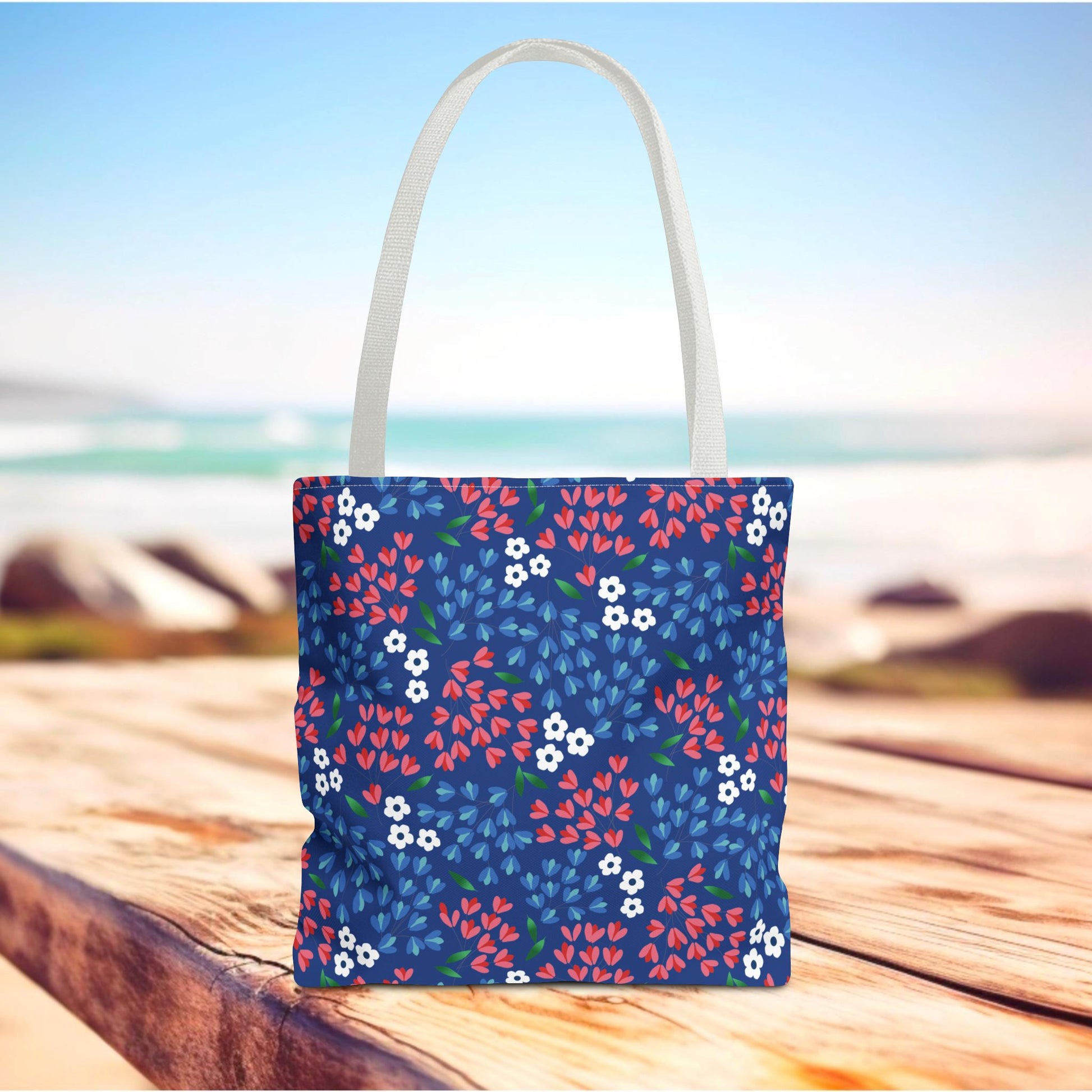 Berry Blue Festival Tote Bag stands on a wooden surface, showcasing a vibrant floral pattern with red, white, and blue blossoms against navy. White handles and a serene water backdrop complete this cheerful summer accessory.